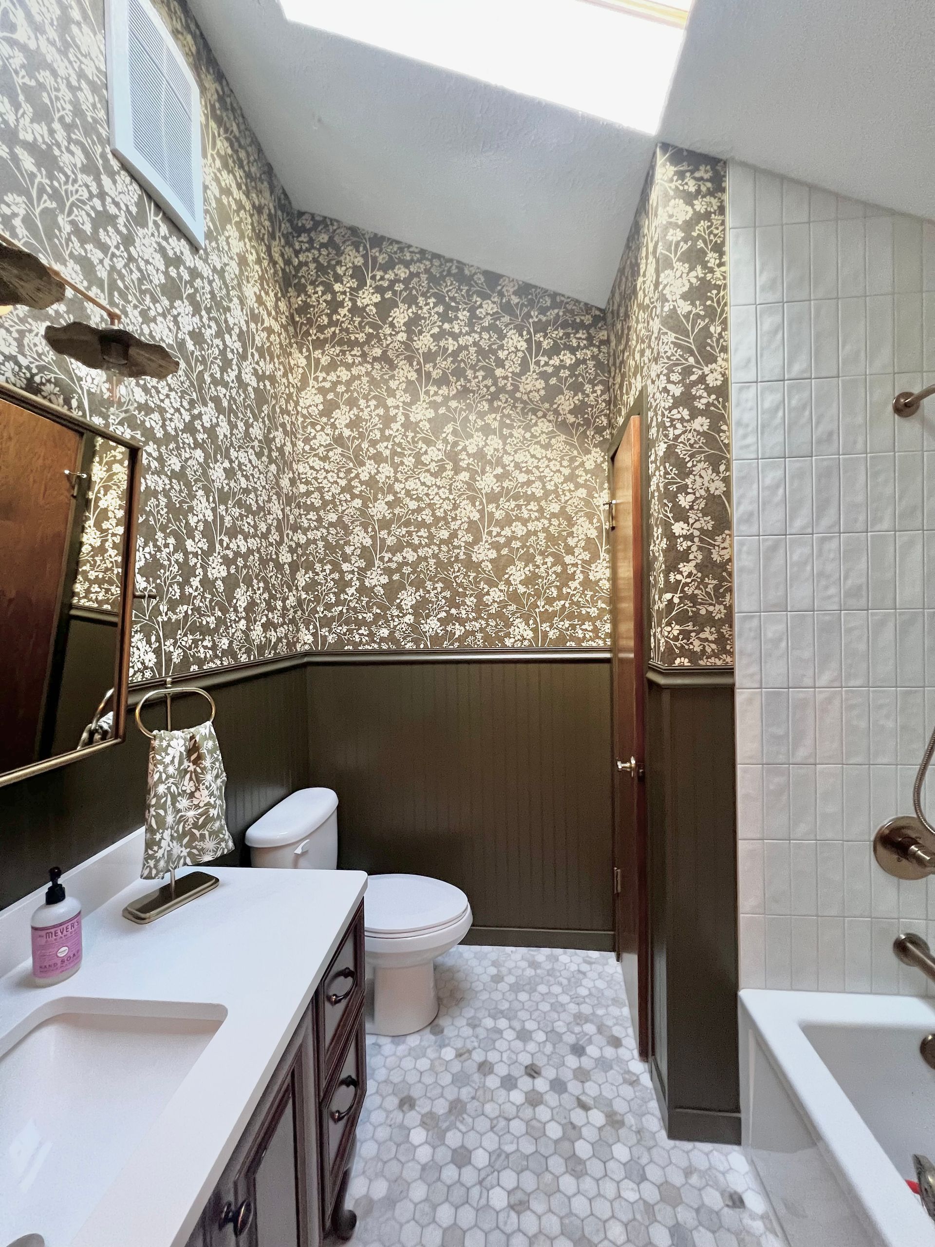 A bathroom with brown and white floral wallpaper above dark wainscoting, a vanity, toilet, and white tile shower.