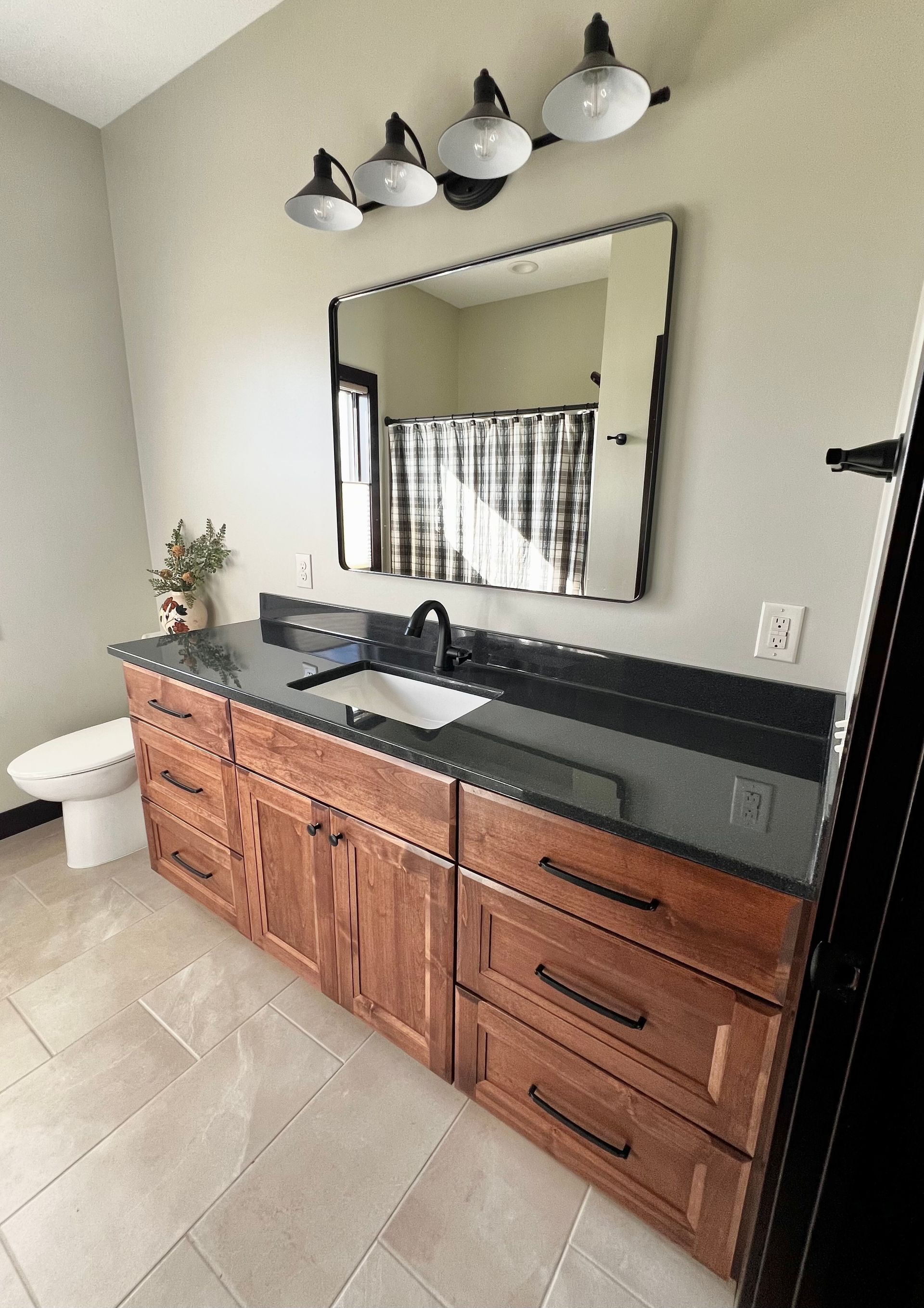 A modern bathroom vanity with wood cabinets, black countertops, a rectangular mirror, and industrial-style lighting.
