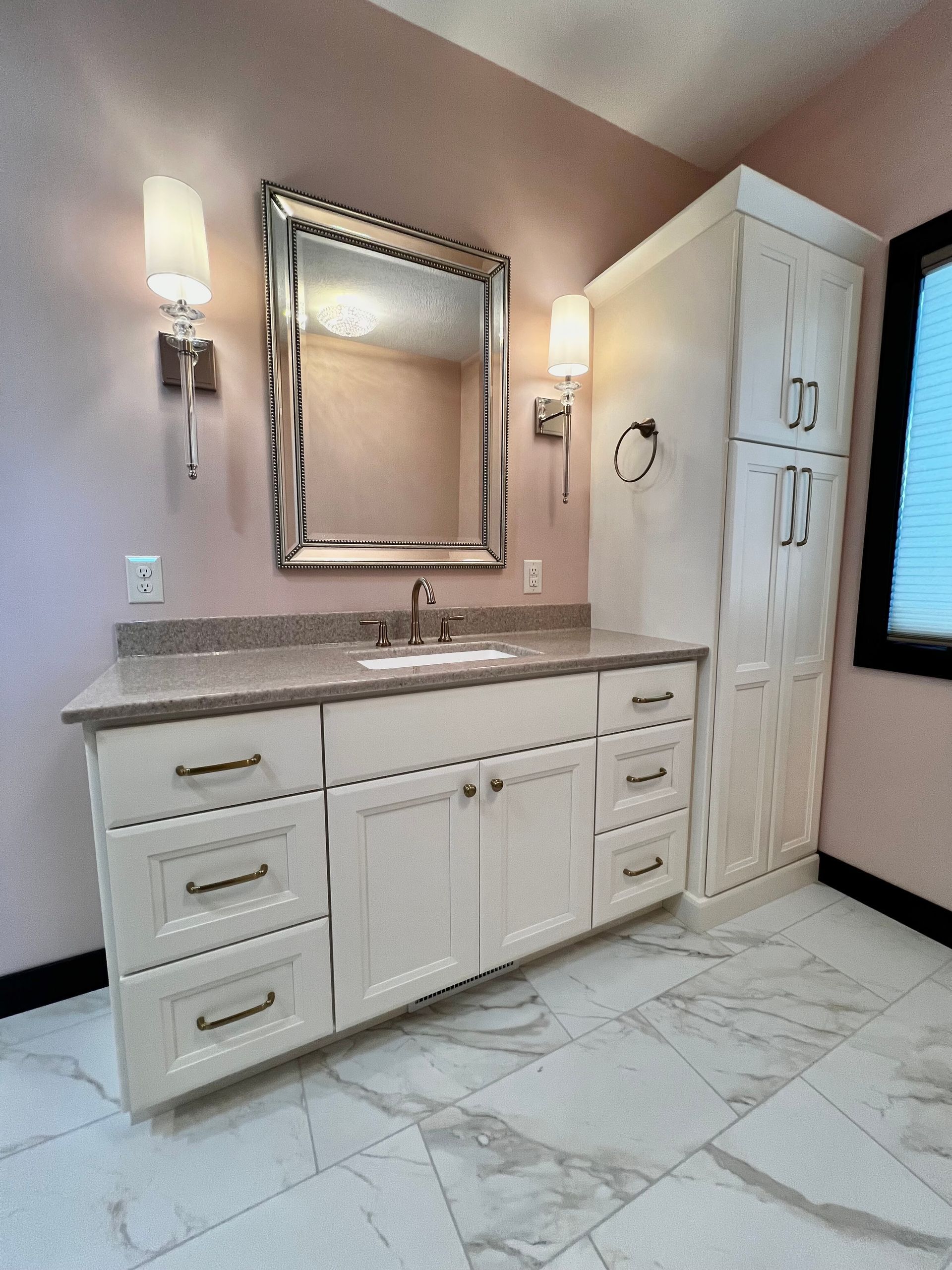 A modern bathroom vanity with white cabinets, a granite countertop, a mirror, and wall sconces on pale pink walls.
