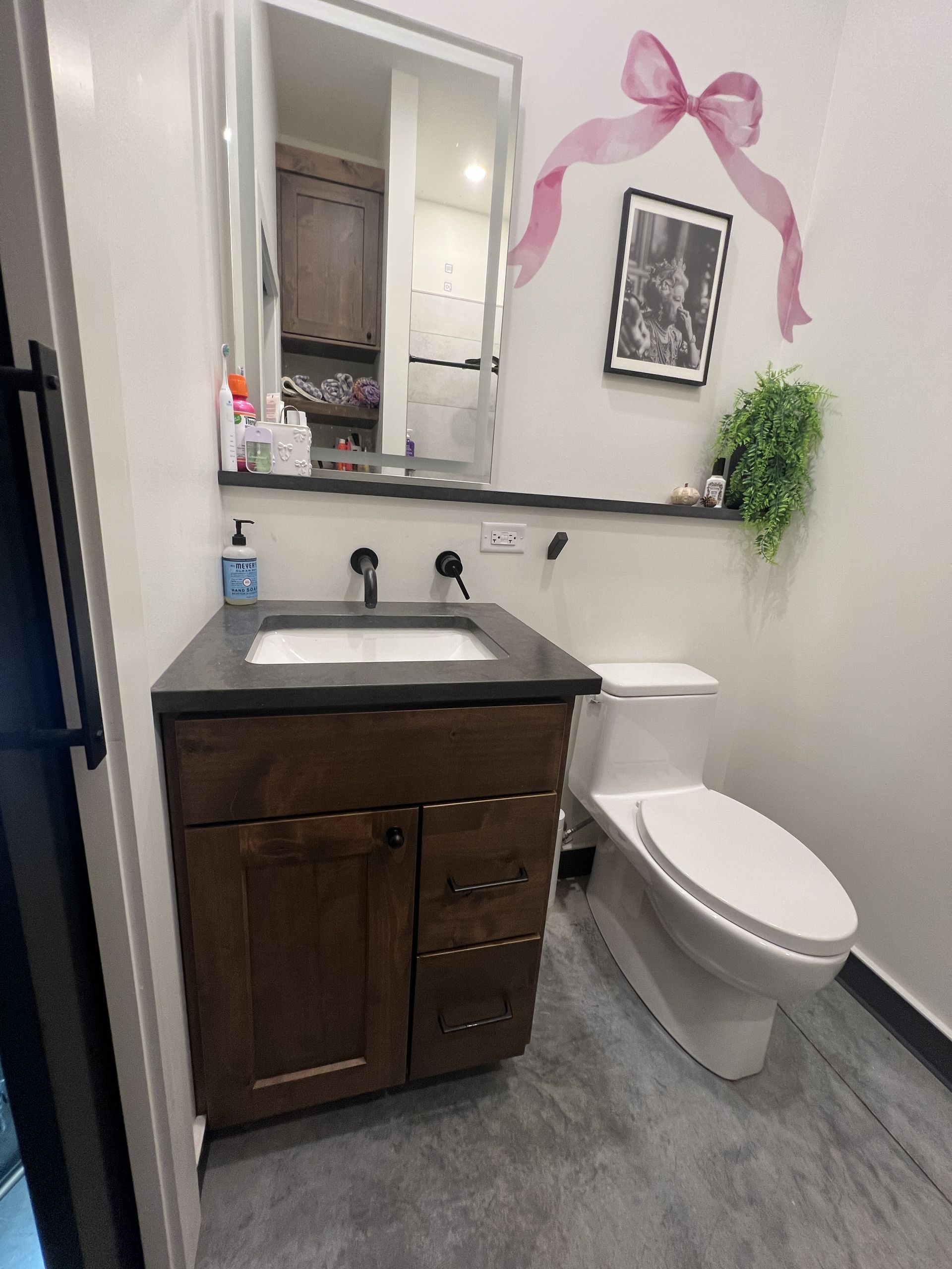 A small bathroom with a wooden vanity, dark countertop, white sink, a mirror, a toilet, and a pink bow decal on the wall.