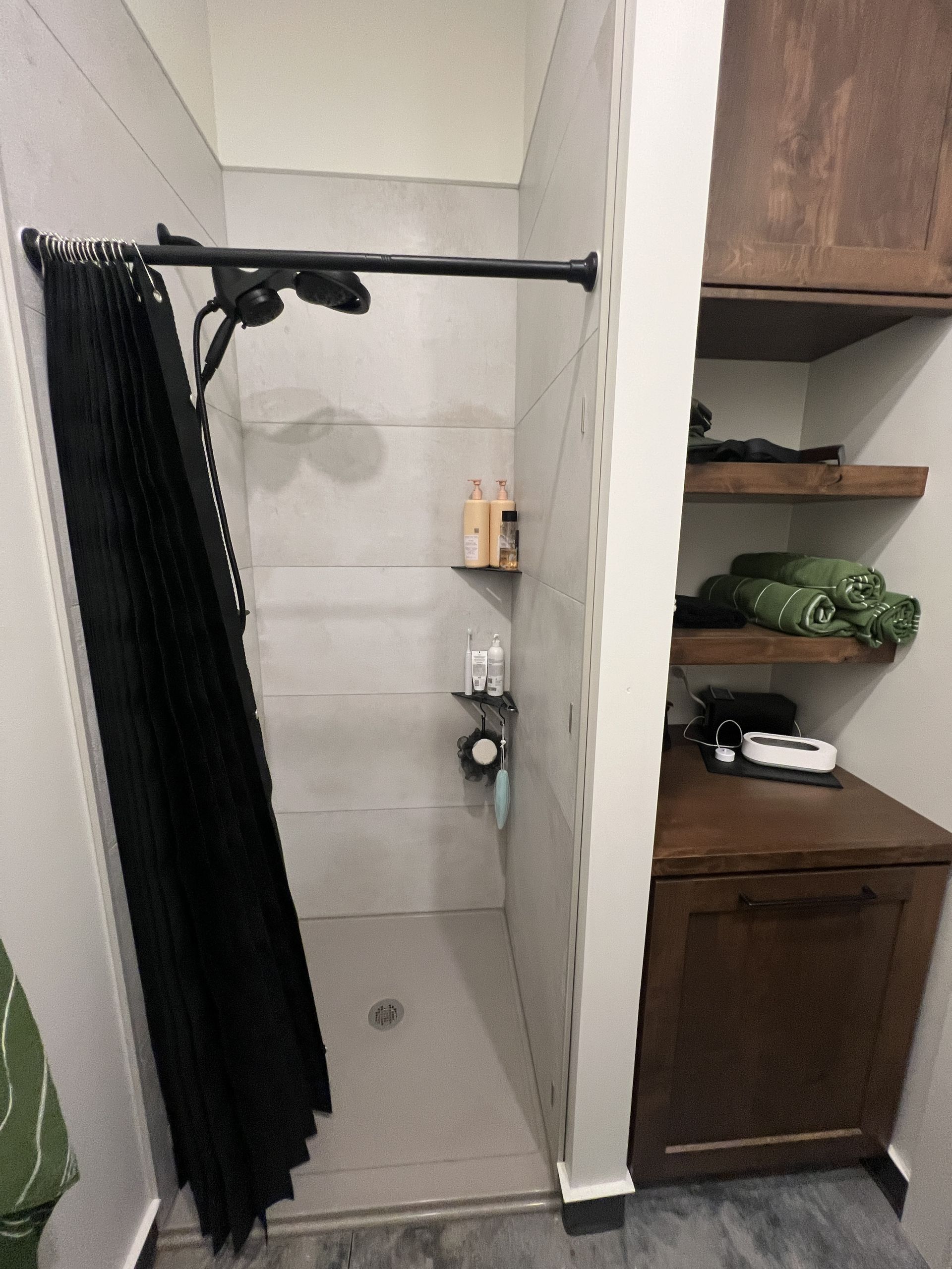 A shower with a black curtain next to built-in wooden shelving and a vanity cabinet in a room with light-colored tile walls.