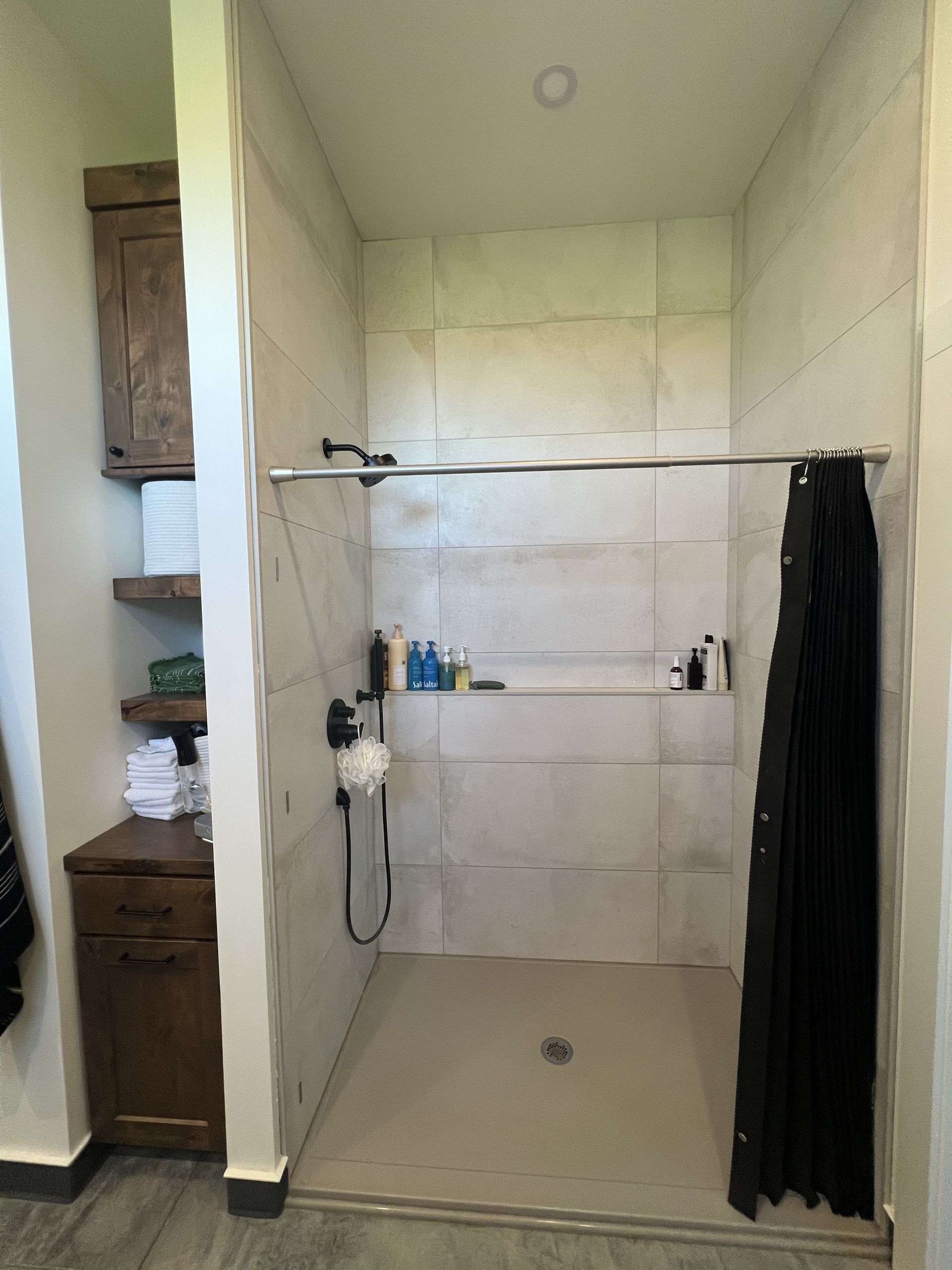 A walk-in shower with large light-colored tiles, a built-in niche, and a black curtain, adjacent to wooden storage shelving.