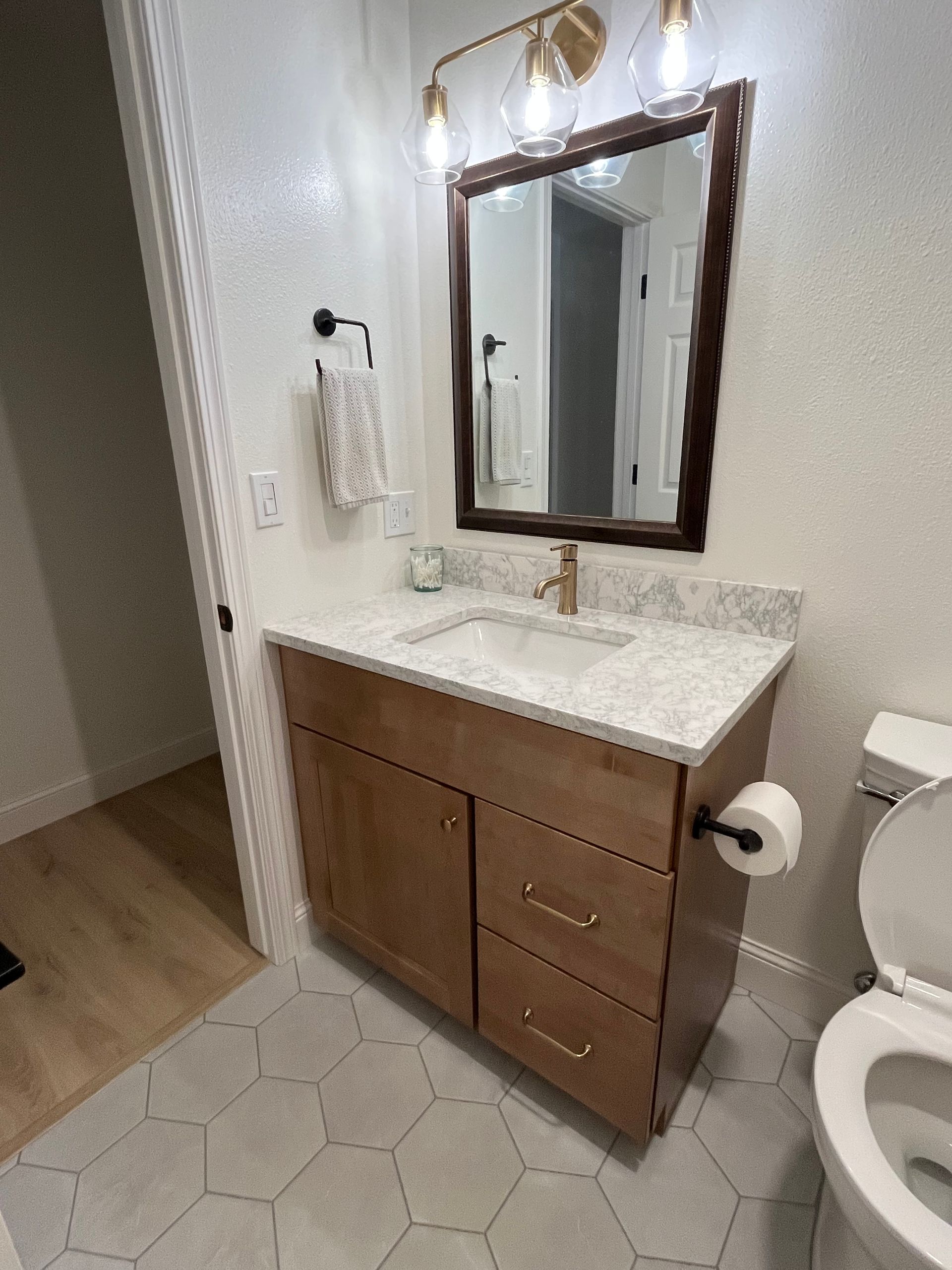 A modern bathroom vanity with a wood cabinet, white stone countertop, rectangular mirror, and gold fixtures on gray tile.