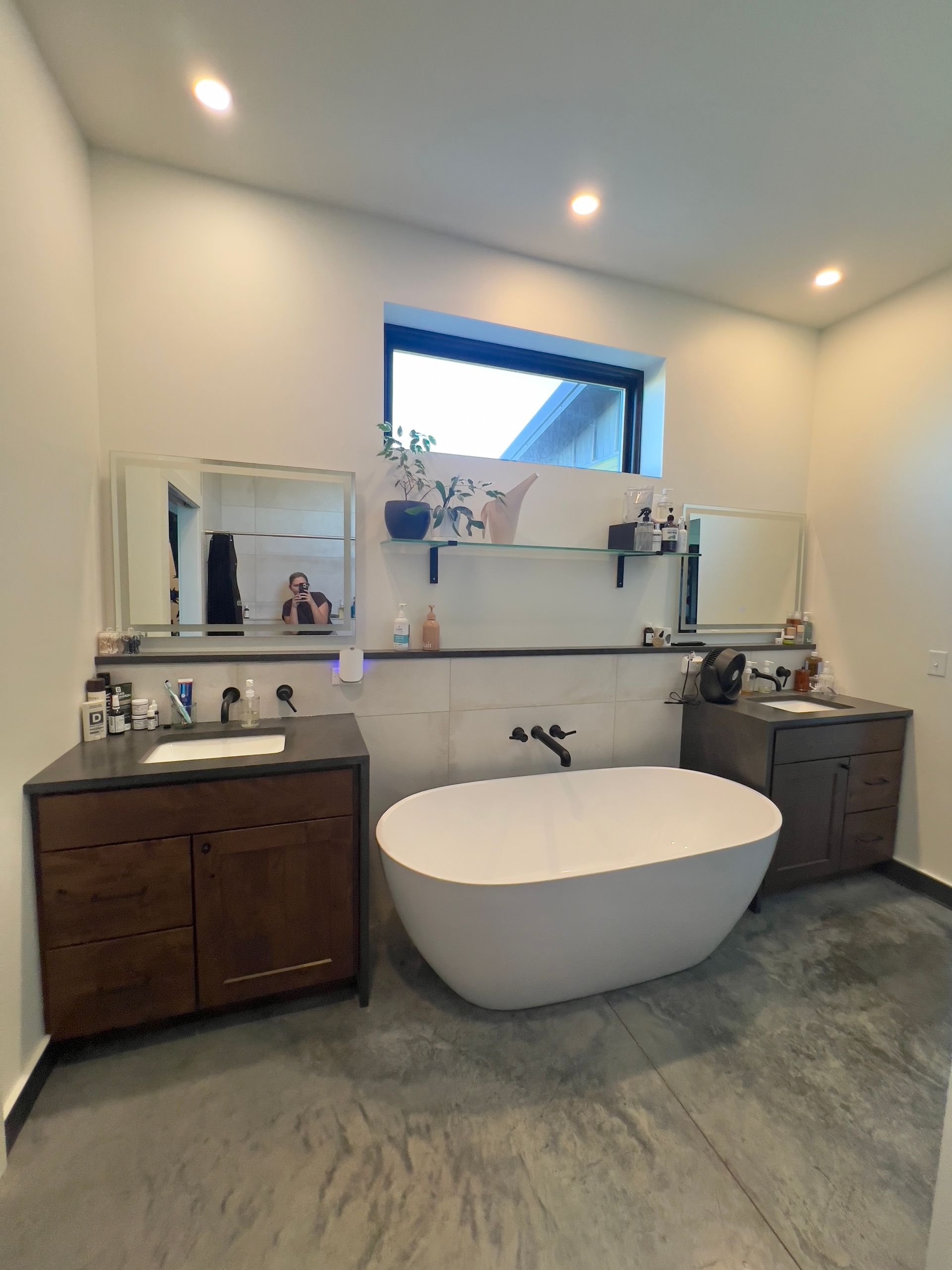 A modern bathroom featuring two dark wood vanities on either side of a white soaking tub beneath a central window.