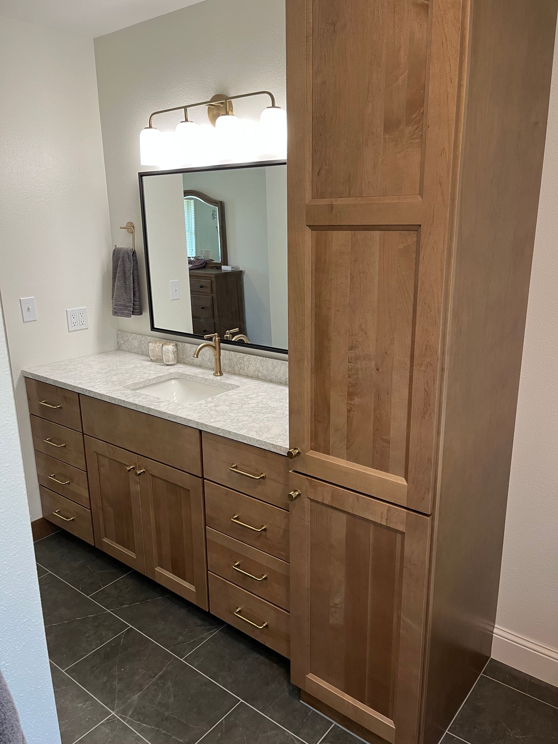 Bathroom vanity with light wood cabinetry, white stone countertop, brass faucet, rectangular mirror, and dark tile floor.
