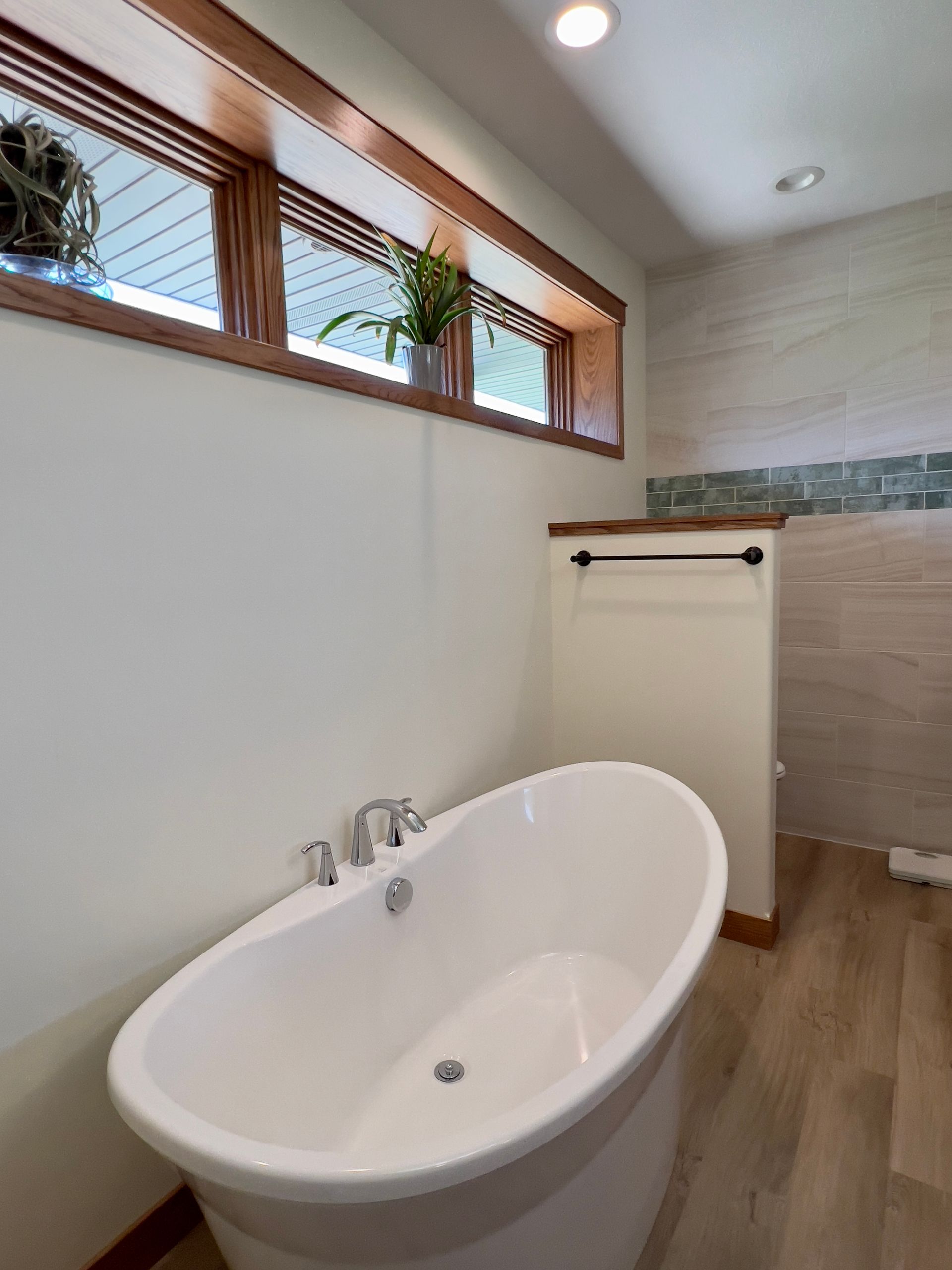 A white freestanding bathtub in a modern bathroom with wood-look flooring, light walls, and a high horizontal window.