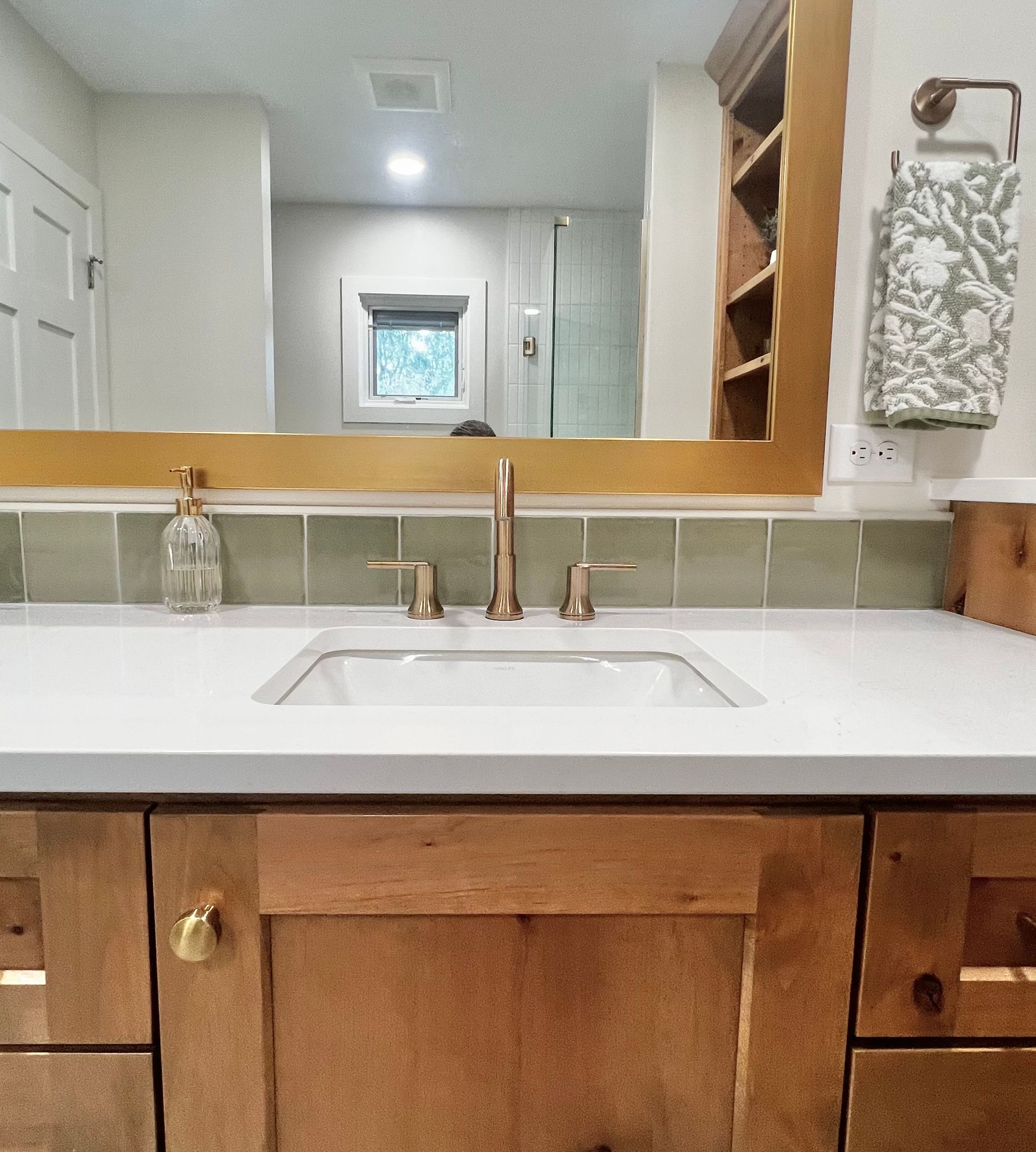A bathroom vanity featuring a white sink, gold faucet, light wood cabinets, green tiled backsplash, and a gold mirror.