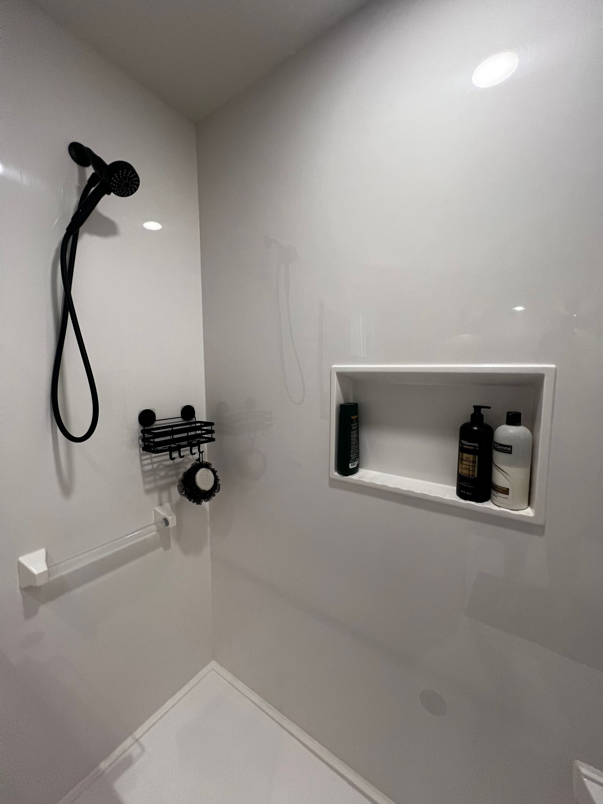A modern walk-in shower with white walls, a black handheld showerhead, wall-mounted bottles, and a recessed wall niche.