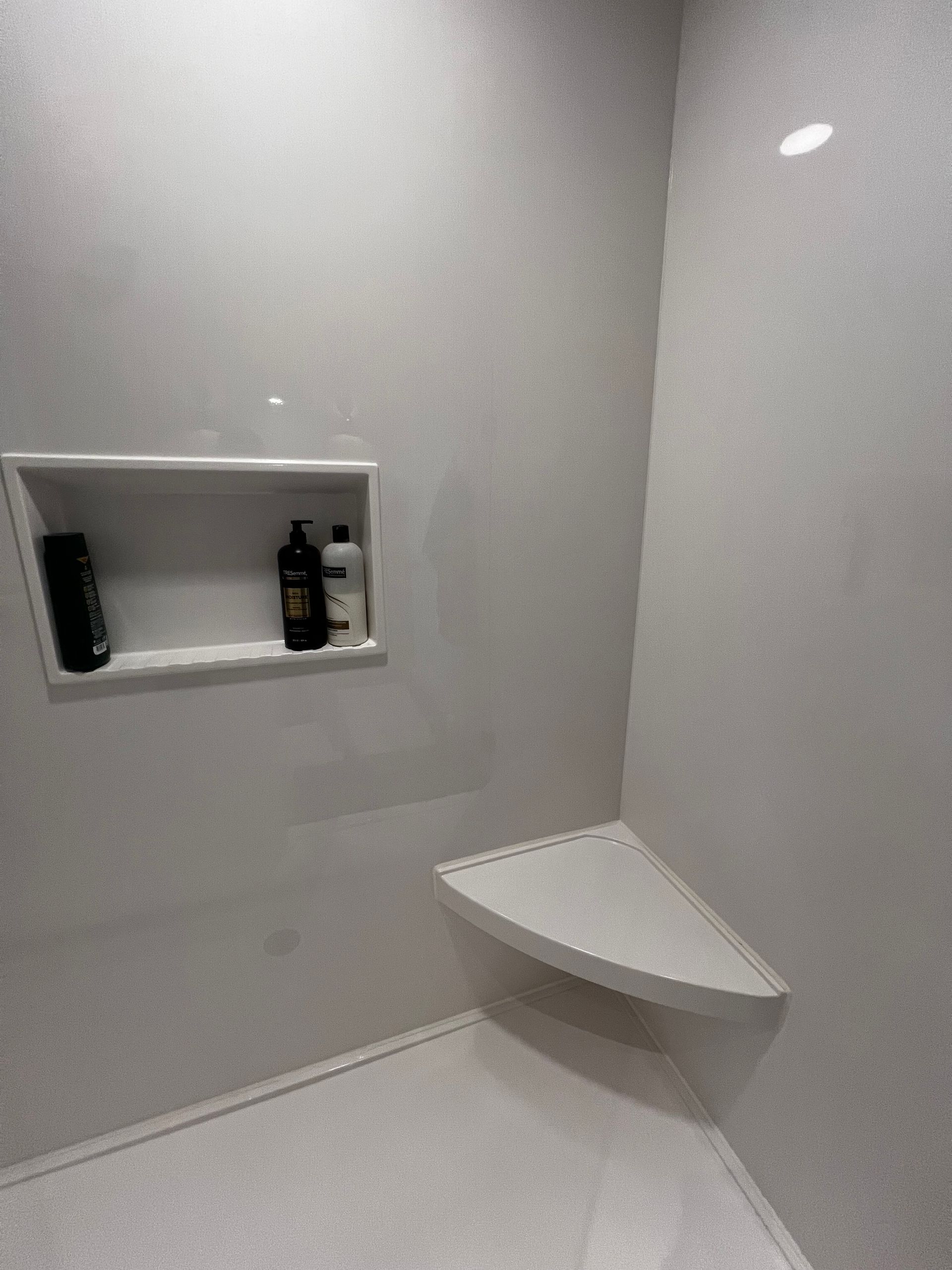A shower interior with light gray walls, a built-in shelf holding two bottles, and a triangular corner seat.