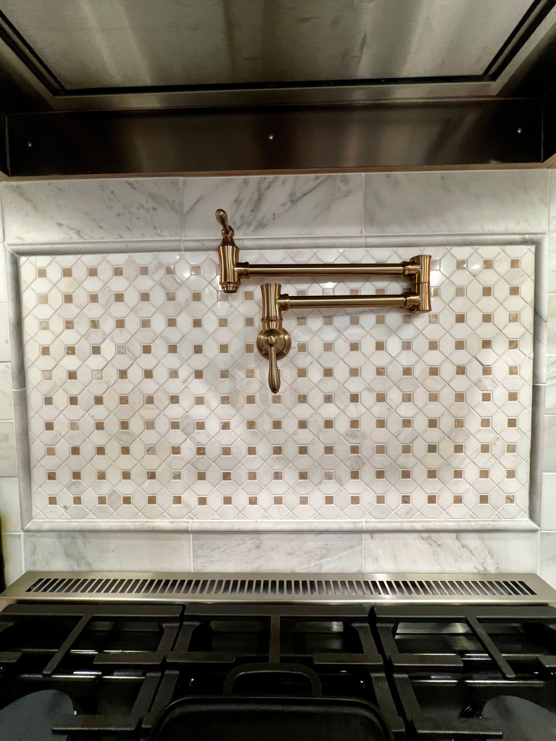 A wall-mounted brass pot filler faucet centered over a kitchen stove with a decorative marble tile backsplash.