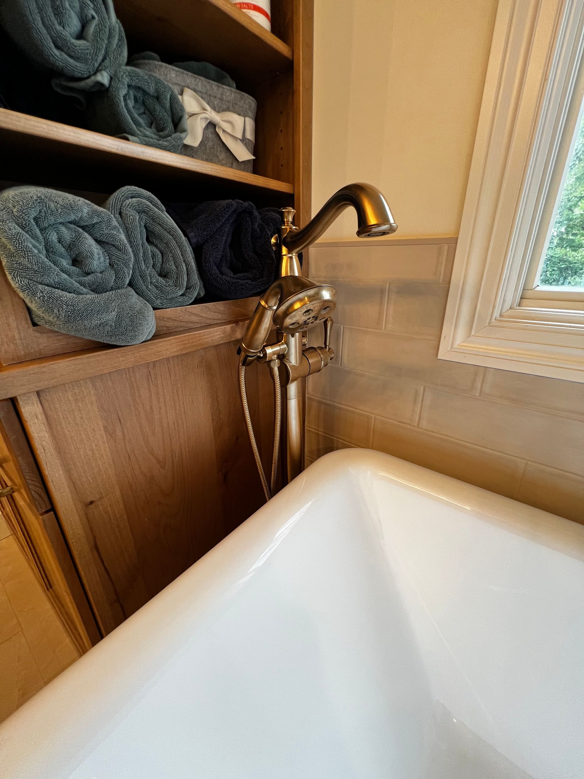 A vintage-style bronze bathtub faucet sits beside a white tub, next to wooden shelves holding rolled grey towels.
