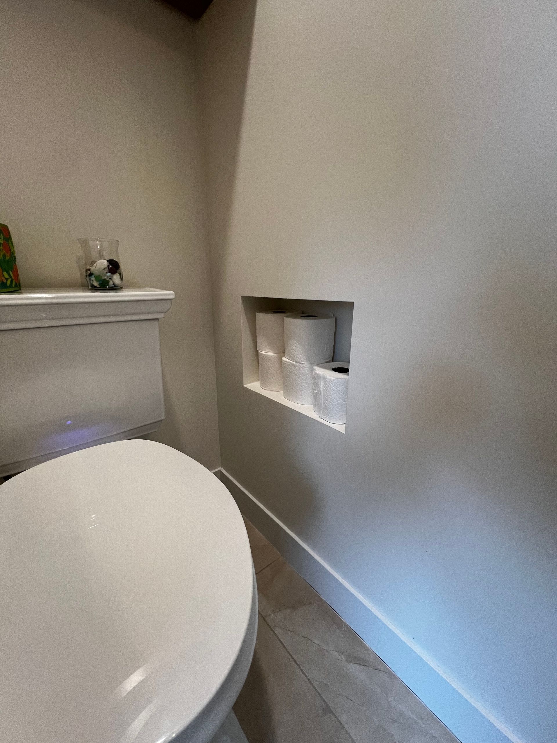 A white toilet in a bathroom with a wall niche containing multiple rolls of toilet paper.