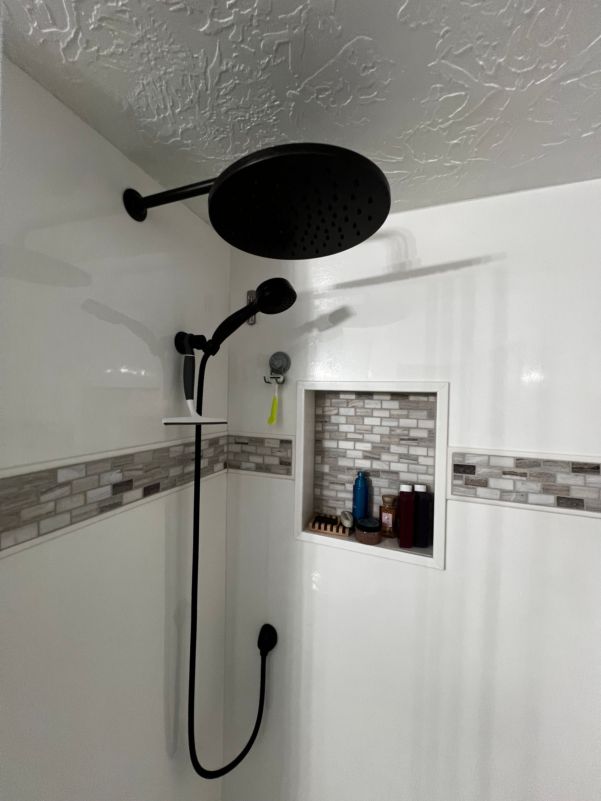 A matte black rainfall and handheld showerhead combo mounted on a white shower wall with a tiled niche for toiletries.