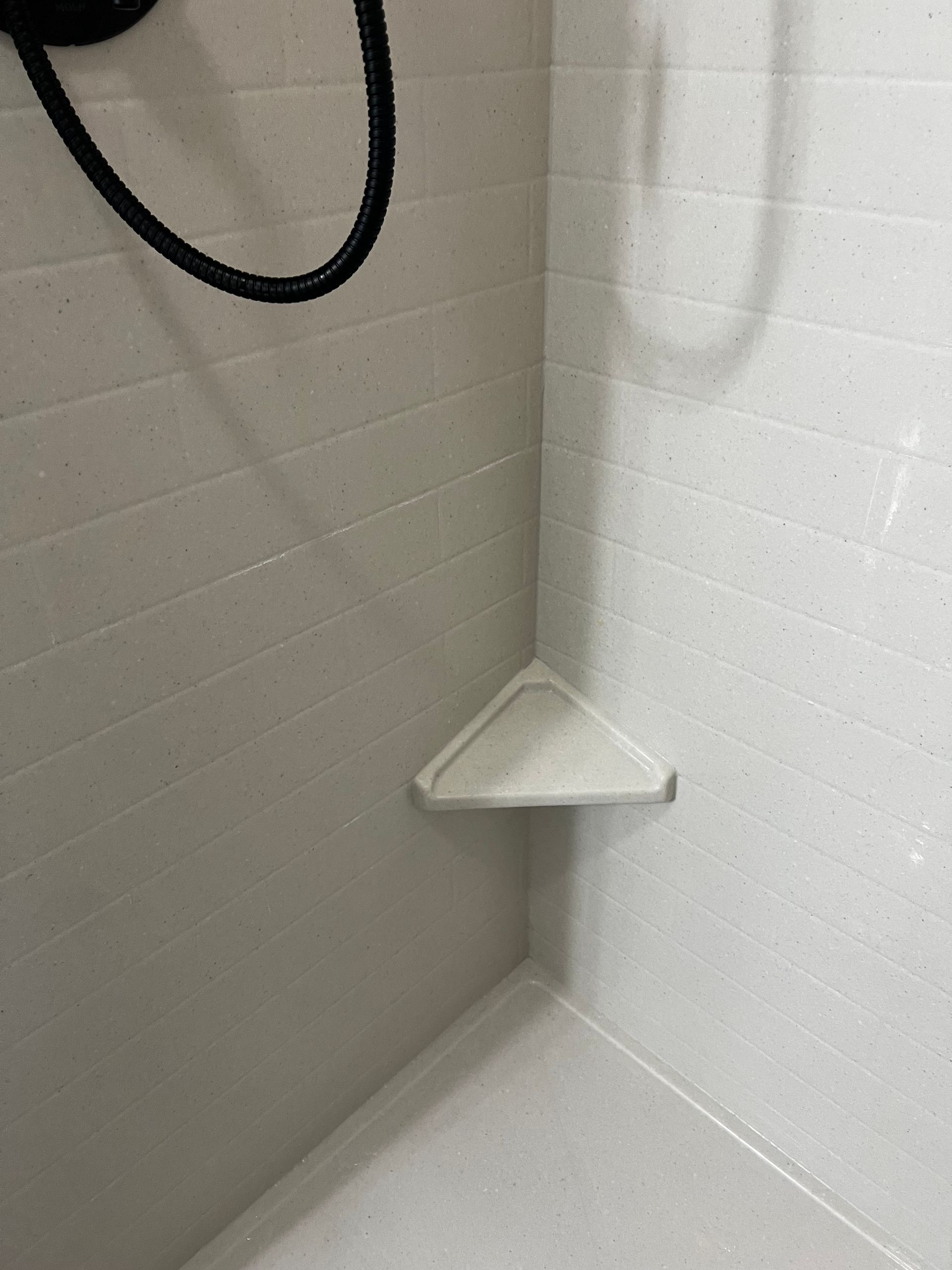 A white triangular corner shelf installed in a white-tiled shower stall.