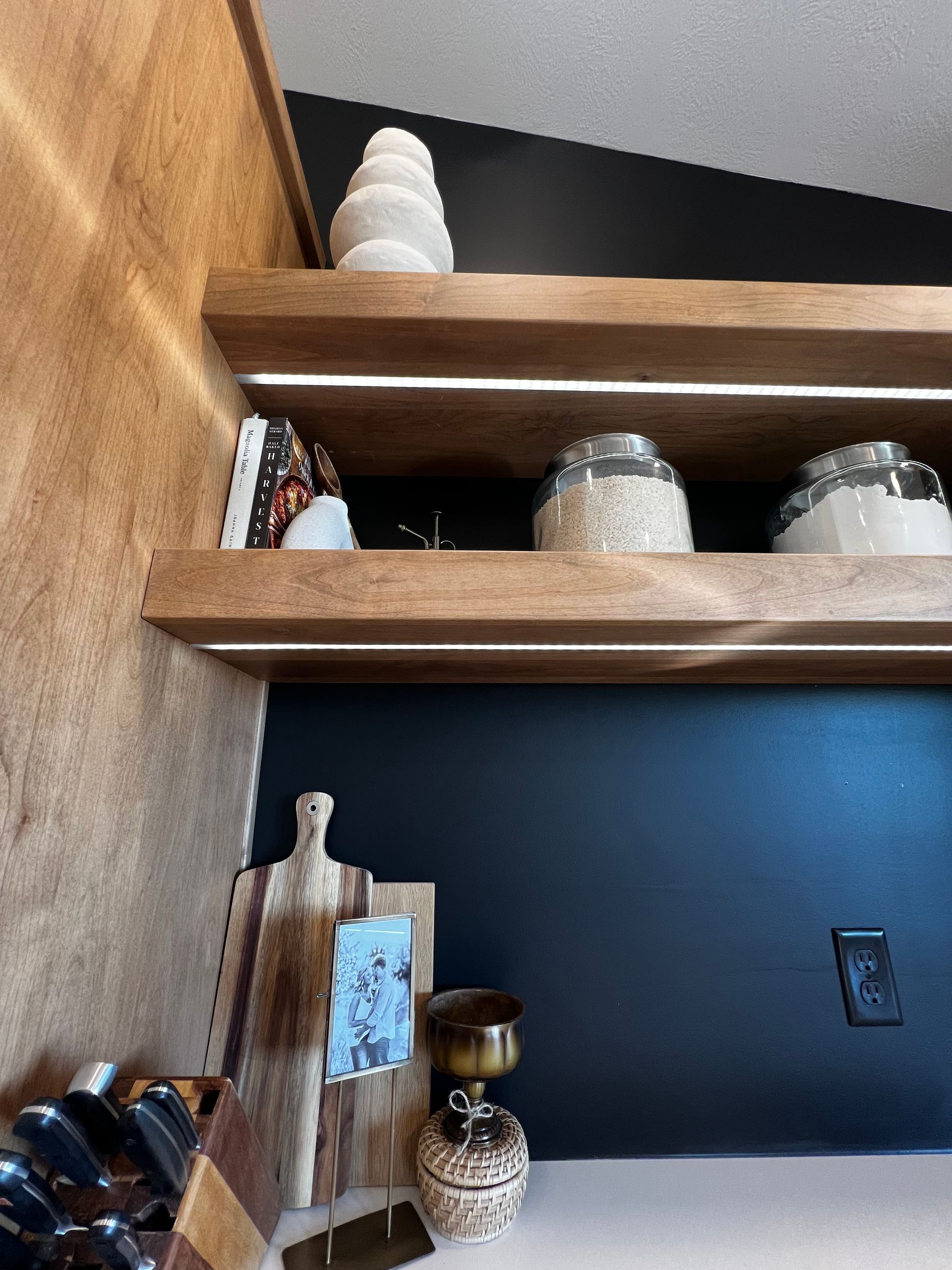 Two wooden floating shelves with integrated lighting above a dark wall, holding decorative jars and kitchen accessories.