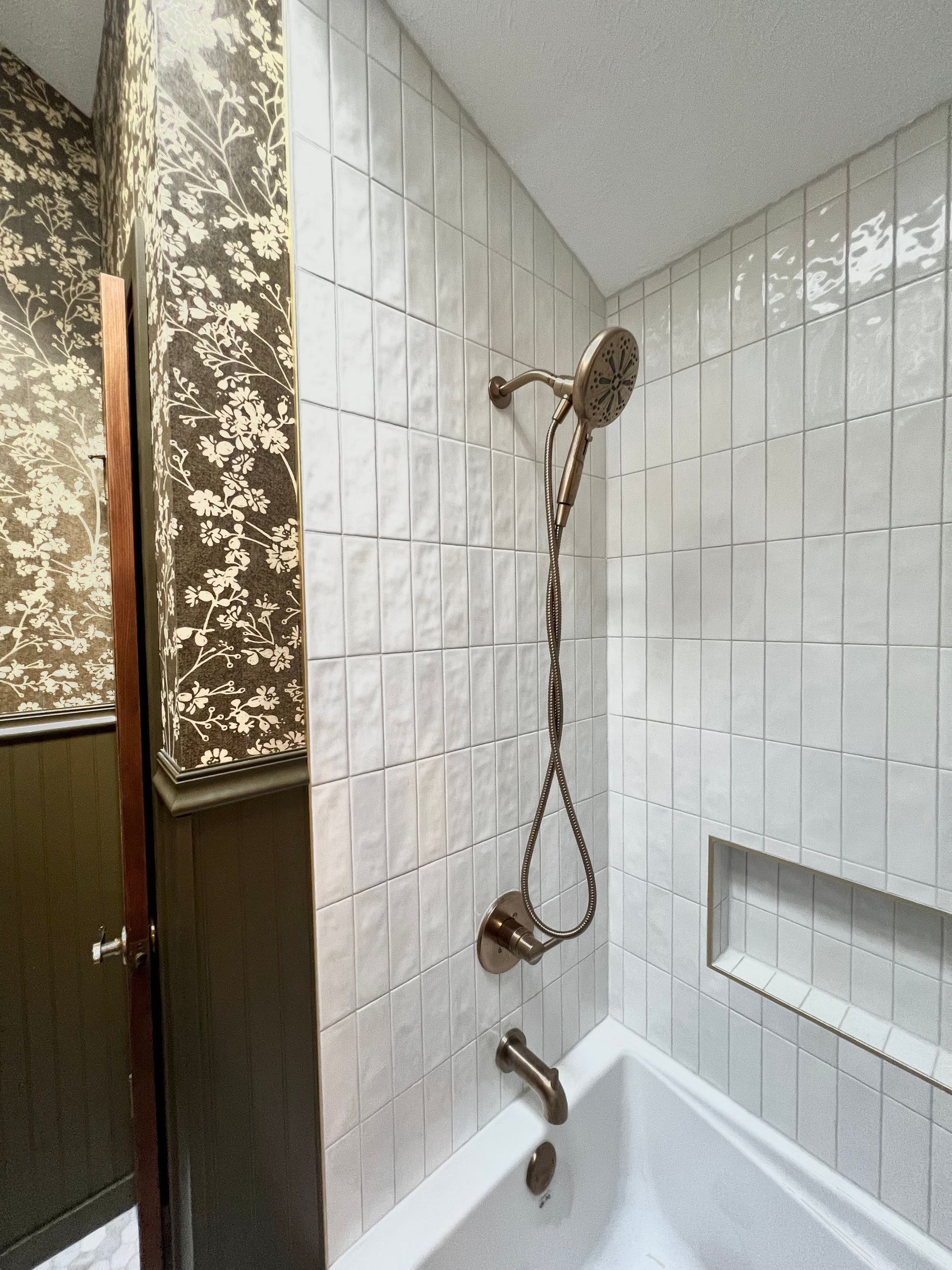 A bathtub and shower area with white vertical tiles, a dark bronze shower fixture, and patterned wallpaper on the wall.