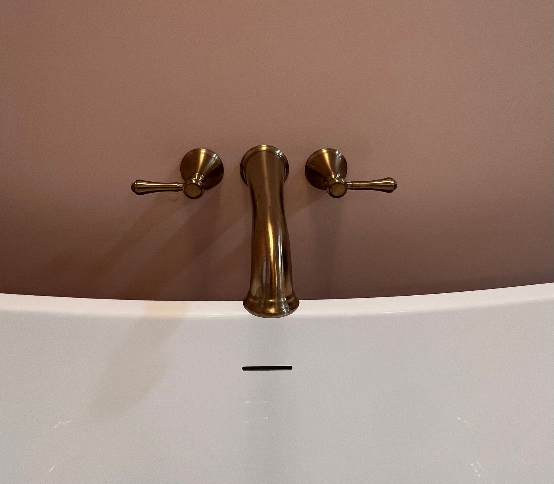 Wall-mounted bronze faucet with two handles centered above a white bathtub against a light pink wall.