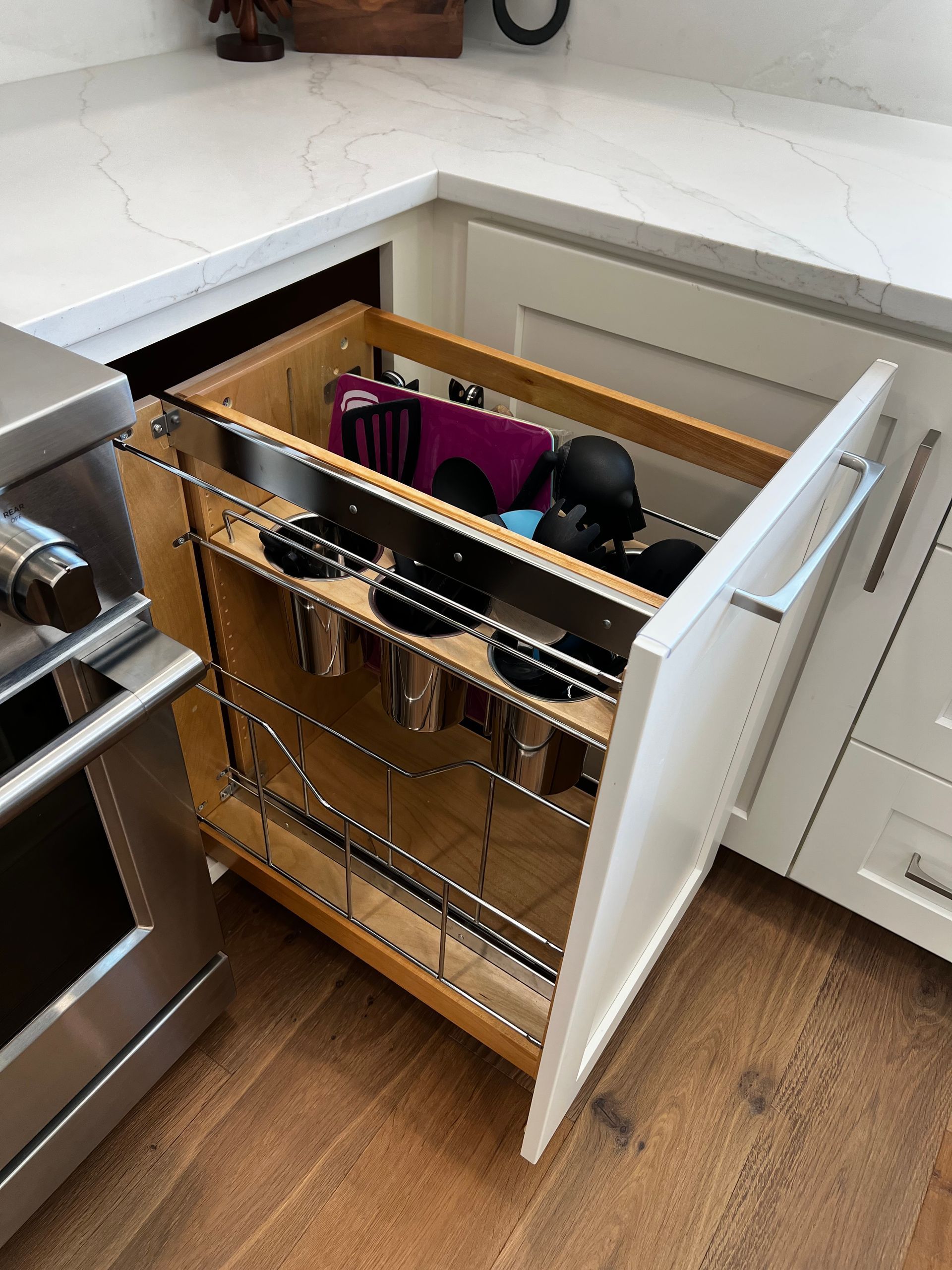 A white kitchen pull-out cabinet drawer containing a chrome utensil organizer next to a stainless steel oven.