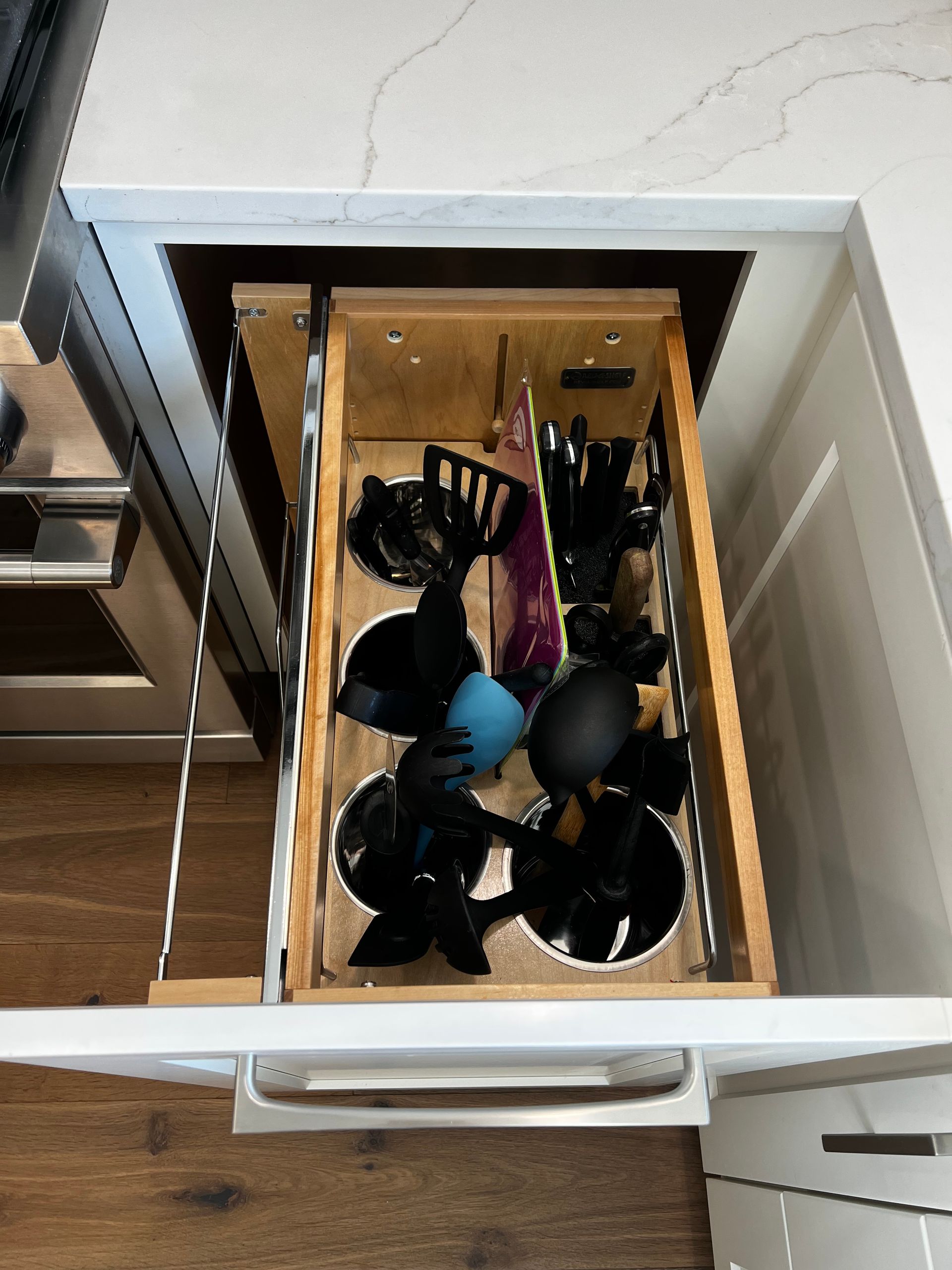 A pull-out kitchen cabinet drawer containing various black cooking utensils, some stored in circular white holders.