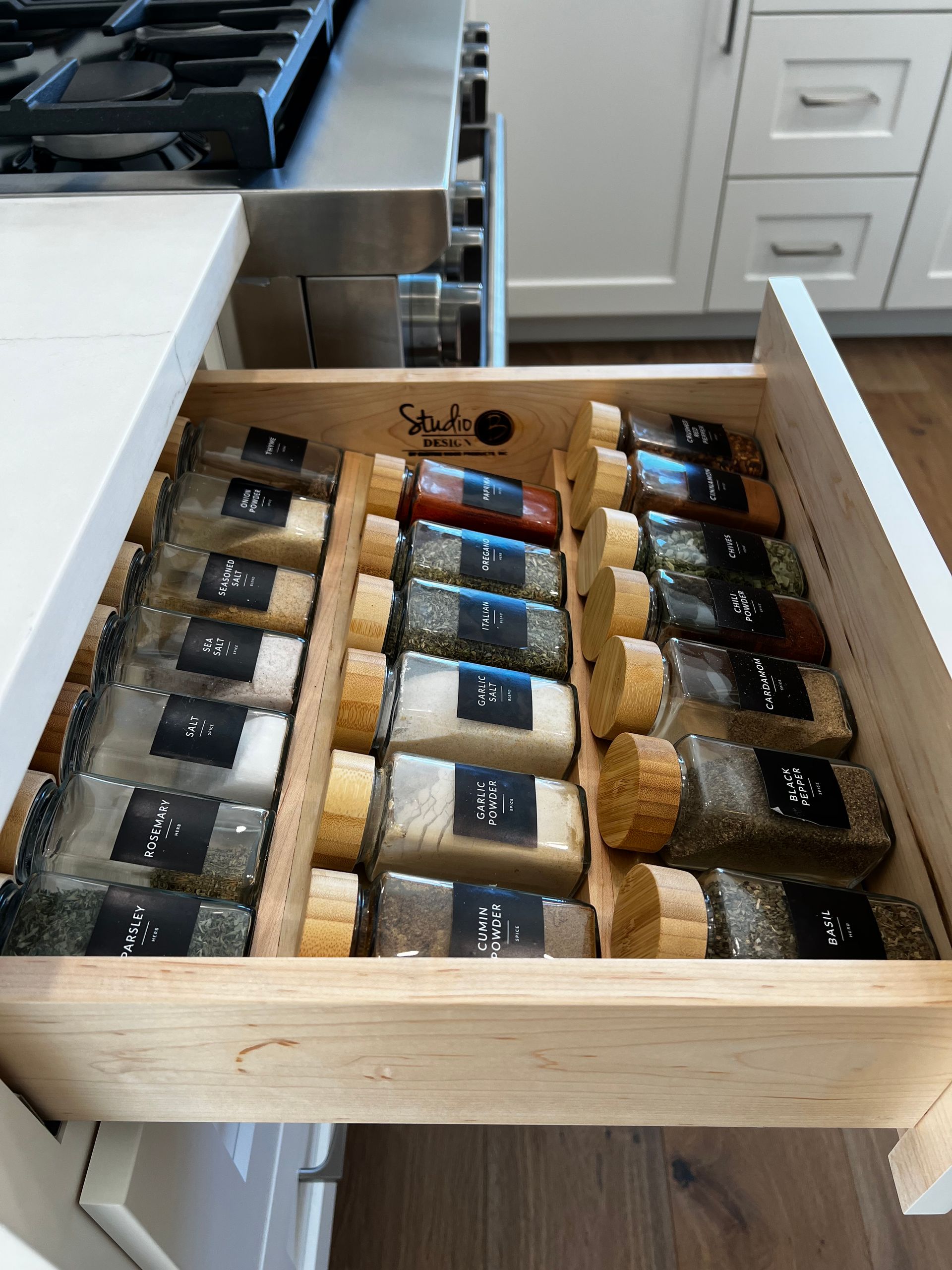 A kitchen drawer pulled open, revealing rows of organized glass spice jars with wooden lids and black labels.