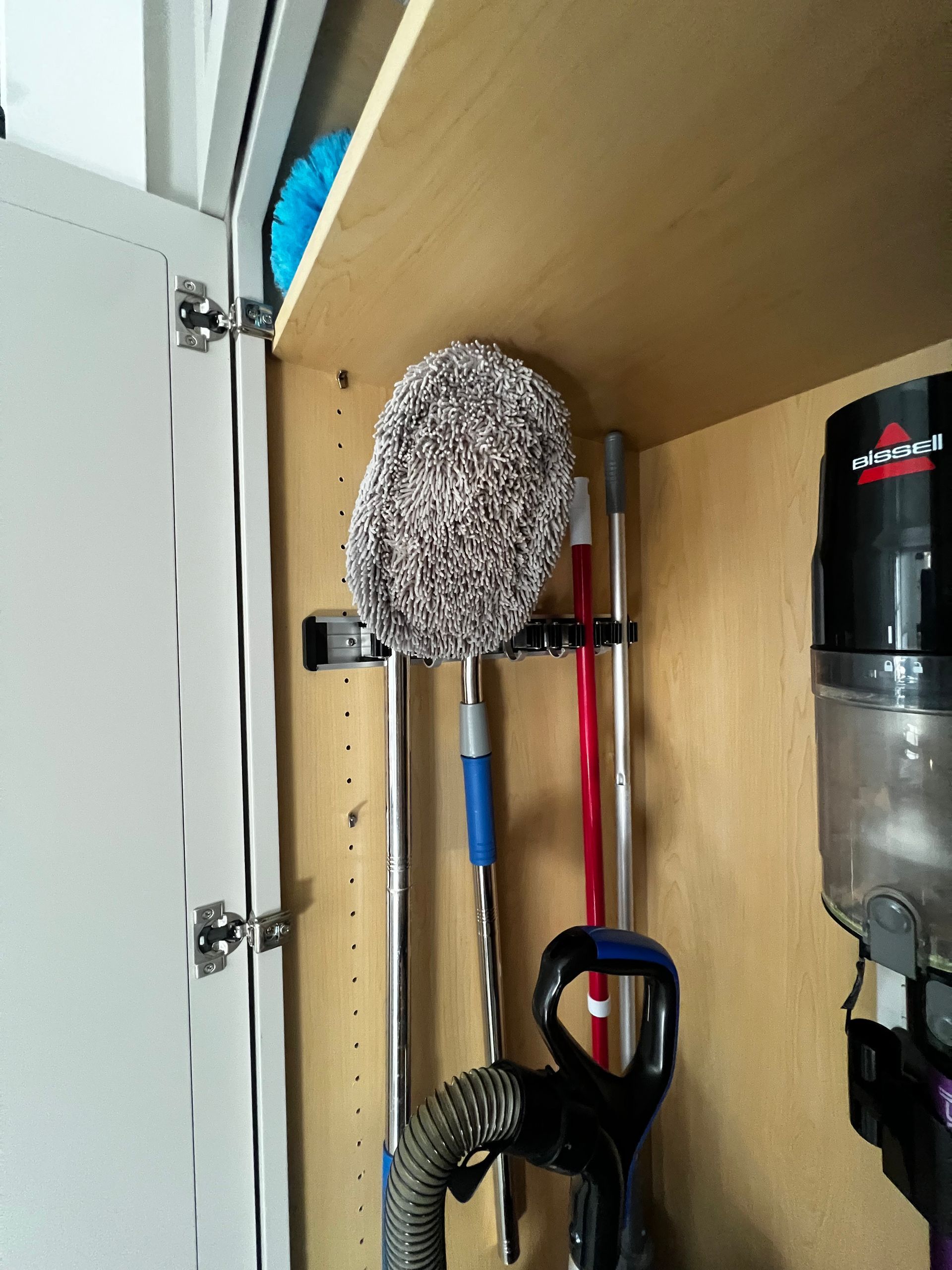 Cleaning tools like mops and a vacuum handle stored on a wall-mounted rack inside a wooden utility cabinet.