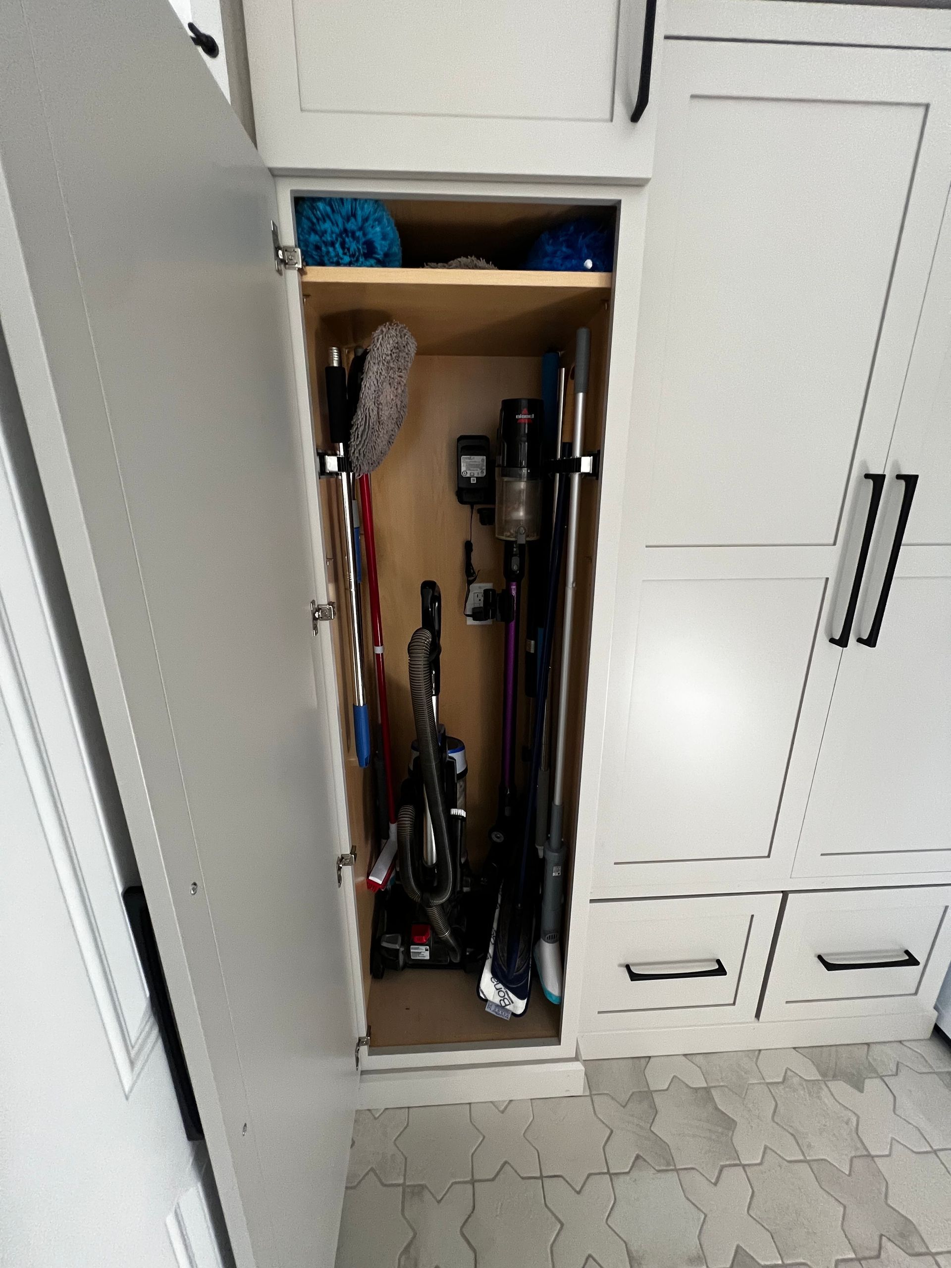 A narrow, white storage cabinet is open, revealing cleaning supplies, including two vacuum cleaners and a mop.