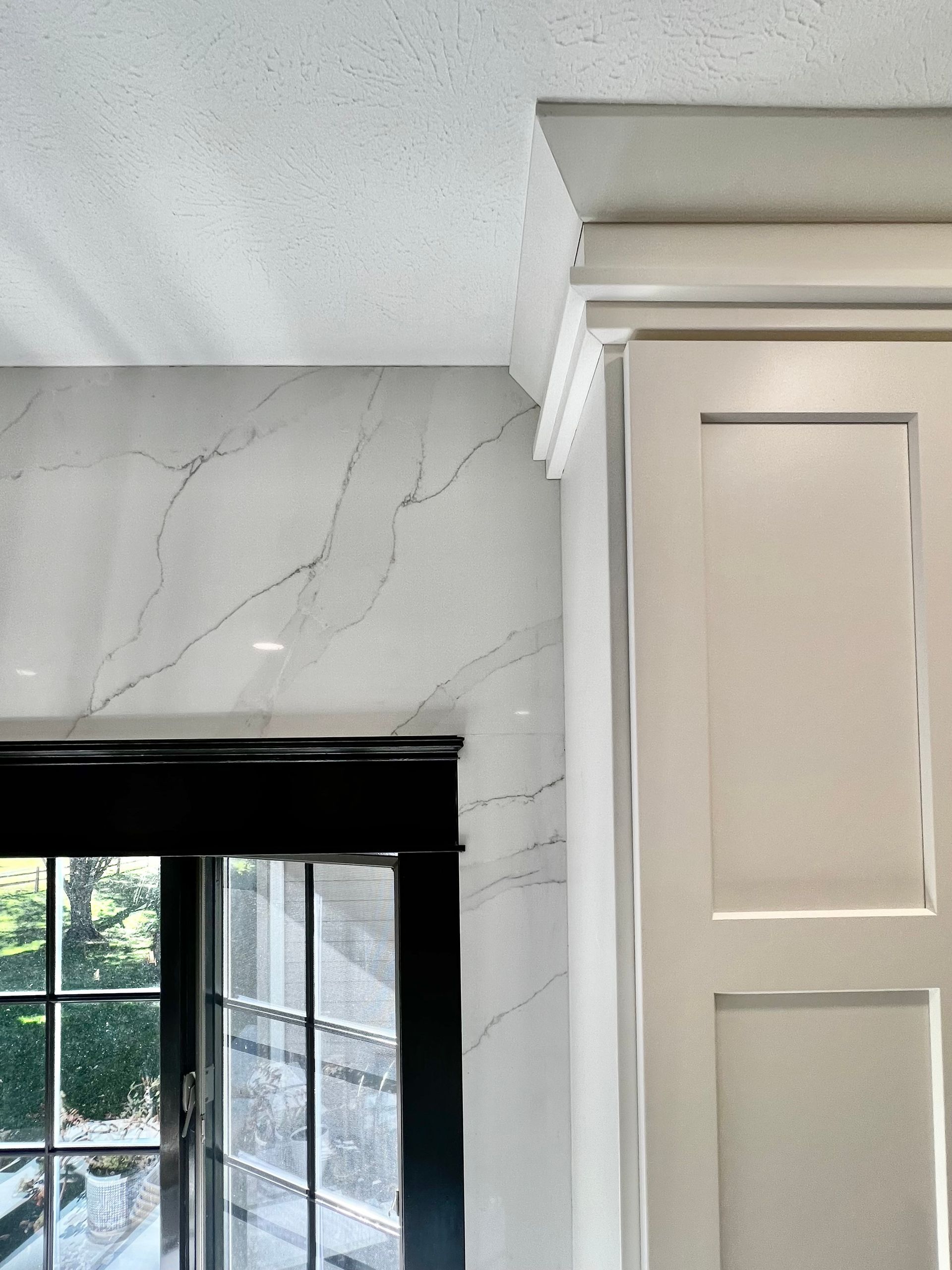 White marble wall with dark veining next to a beige cabinet with crown molding and a window with a black shade.
