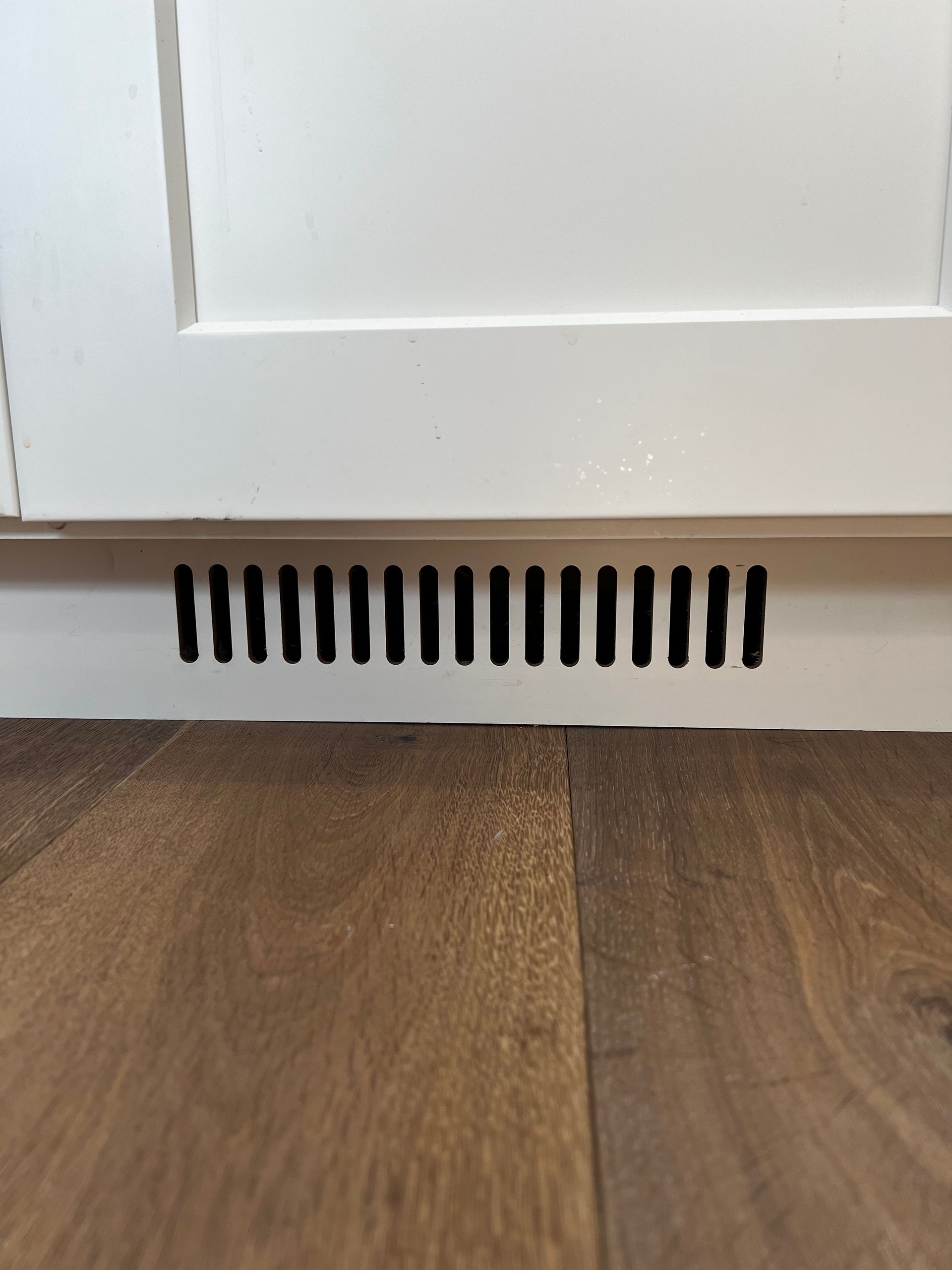 A rectangular vent with vertical slats cut into the white baseboard of a kitchen cabinet above a brown wood-look floor.