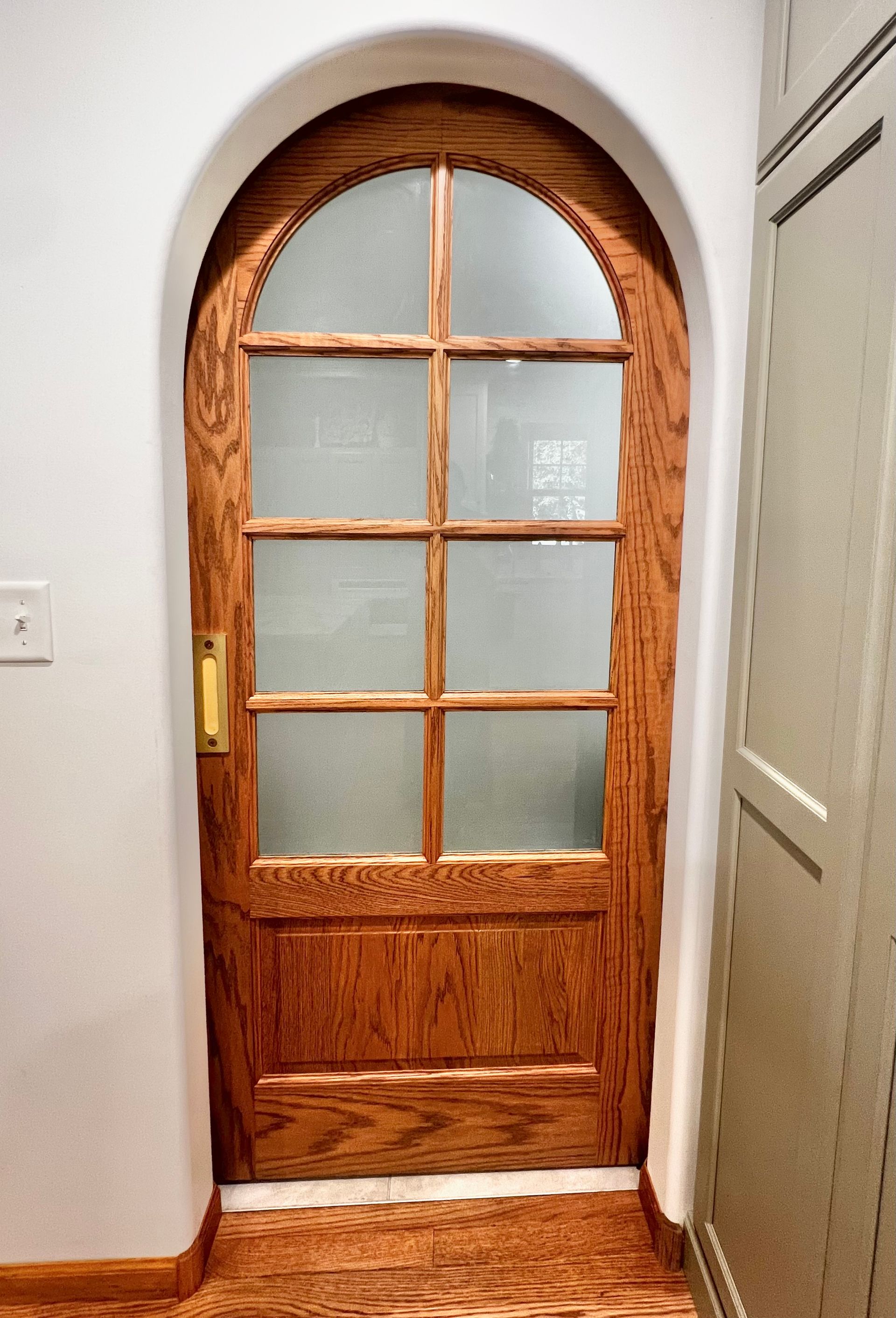 An arched, natural-wood door with six frosted glass panes, set in a white wall above a wooden floor.
