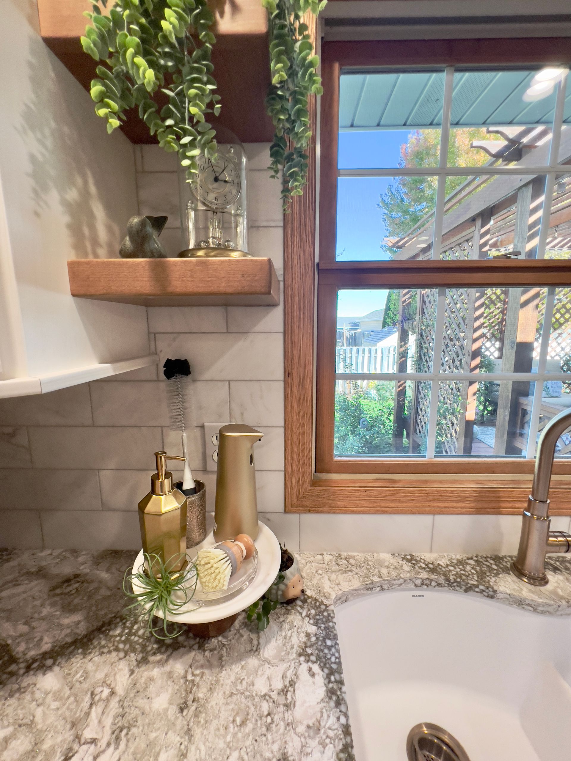 A kitchen sink area featuring a white subway tile backsplash, a wooden shelf with greenery, and gold soap dispensers.