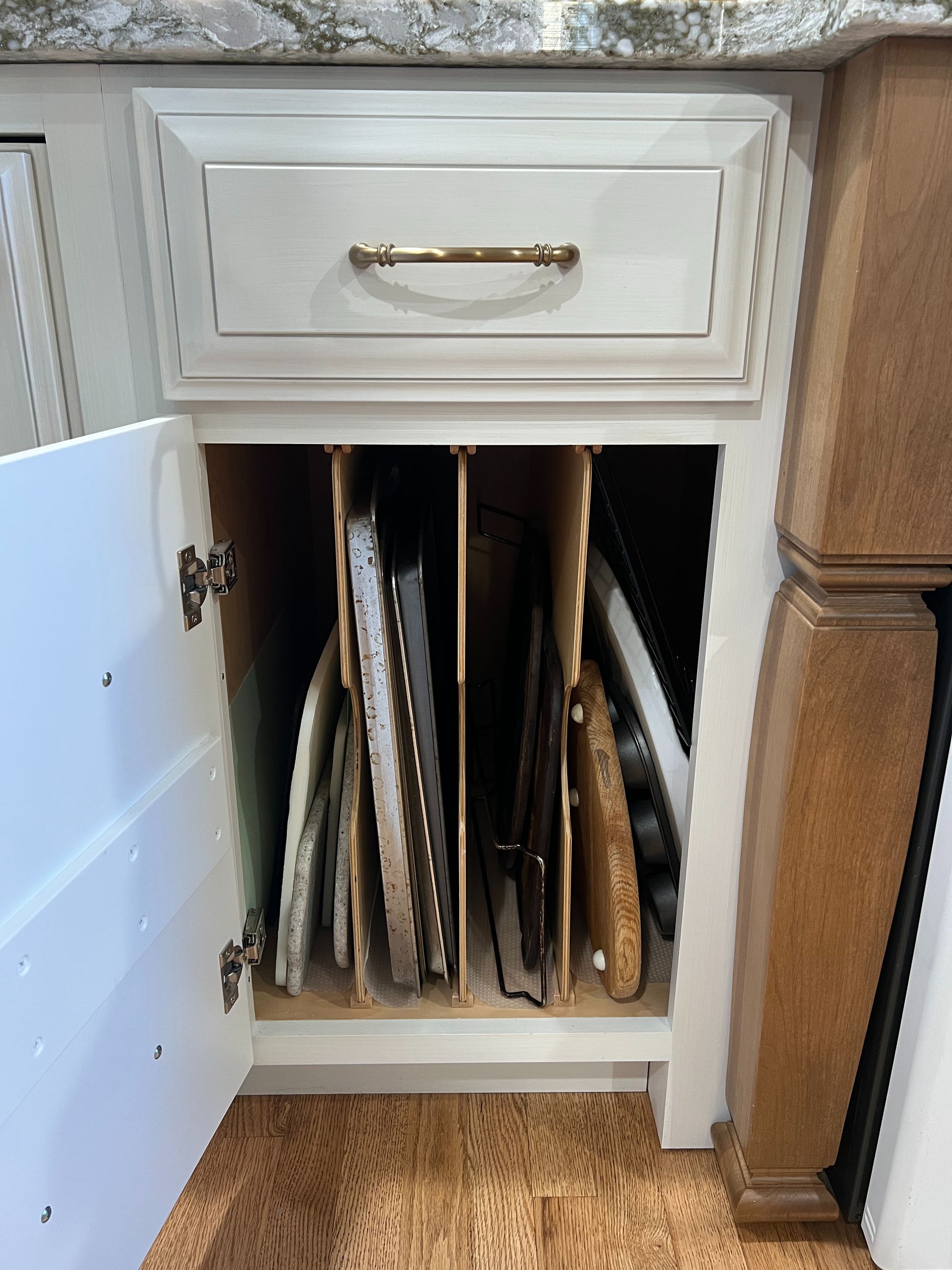 An open white kitchen cabinet with vertical wooden dividers, storing baking sheets and trays upright.