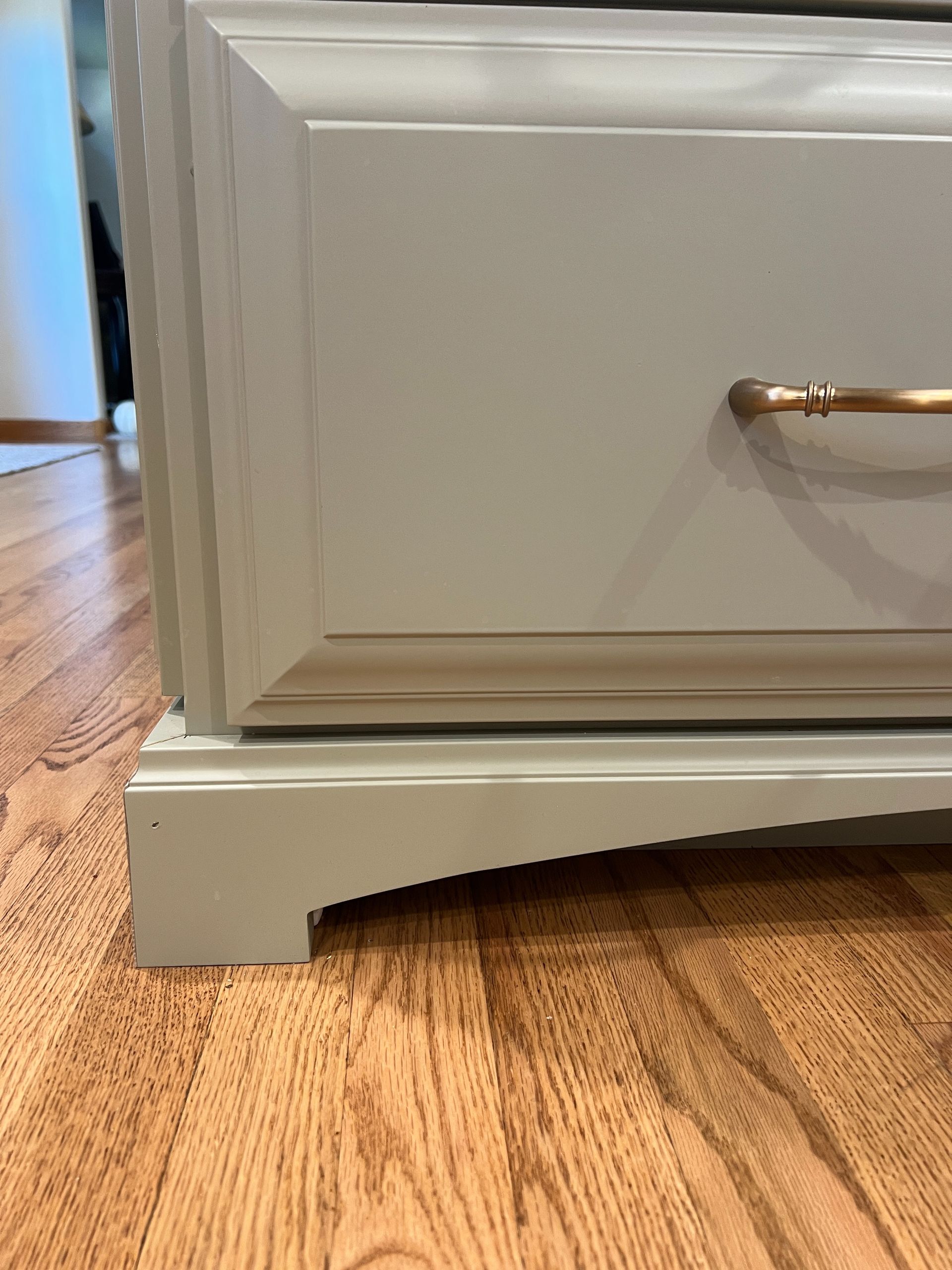 A close-up view of the bottom corner of a light grey painted wooden dresser with a gold handle on a hardwood floor.