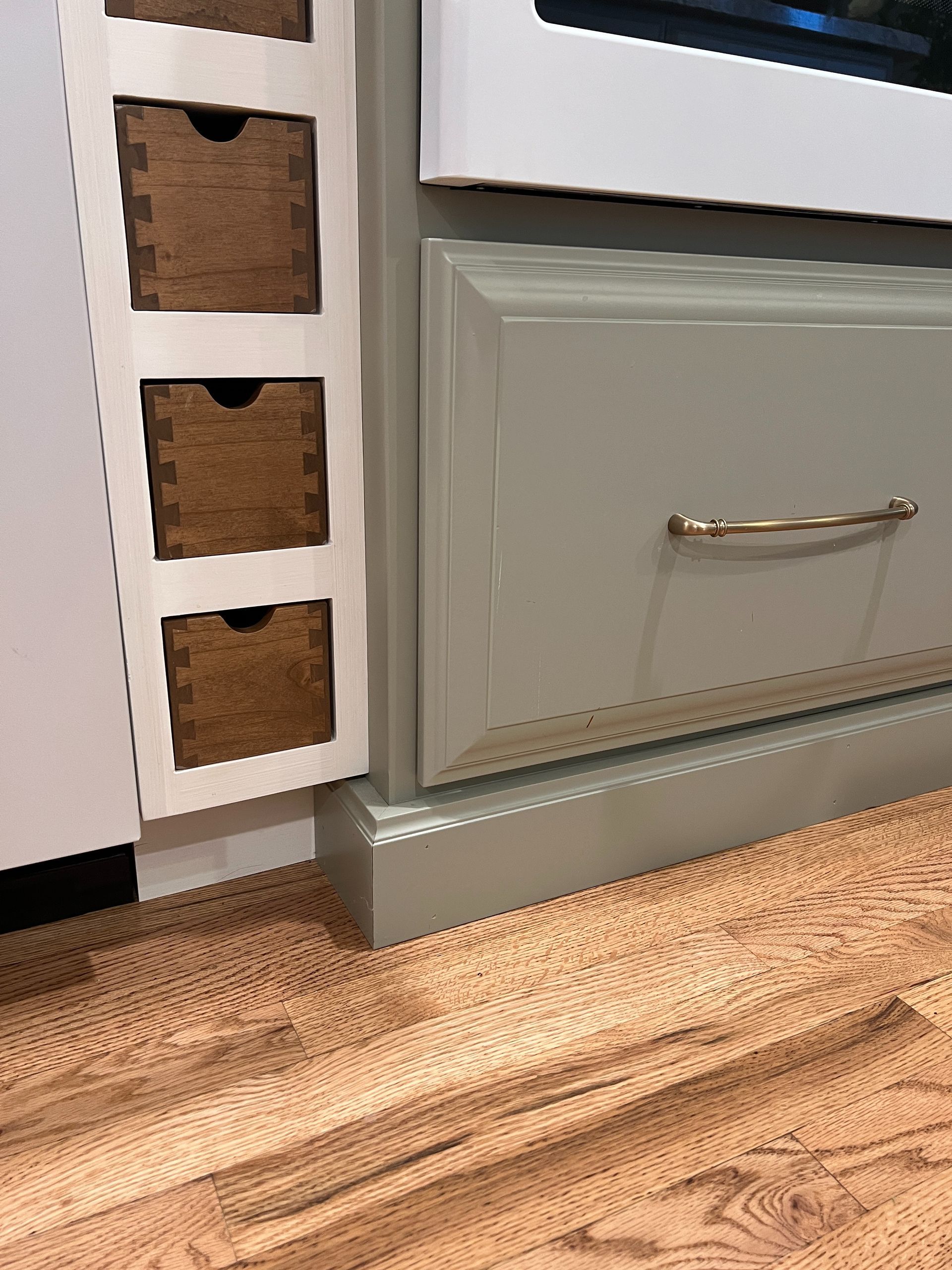 A sage green kitchen cabinet with a brass handle next to a white unit containing three small, wooden open-shelf baskets.