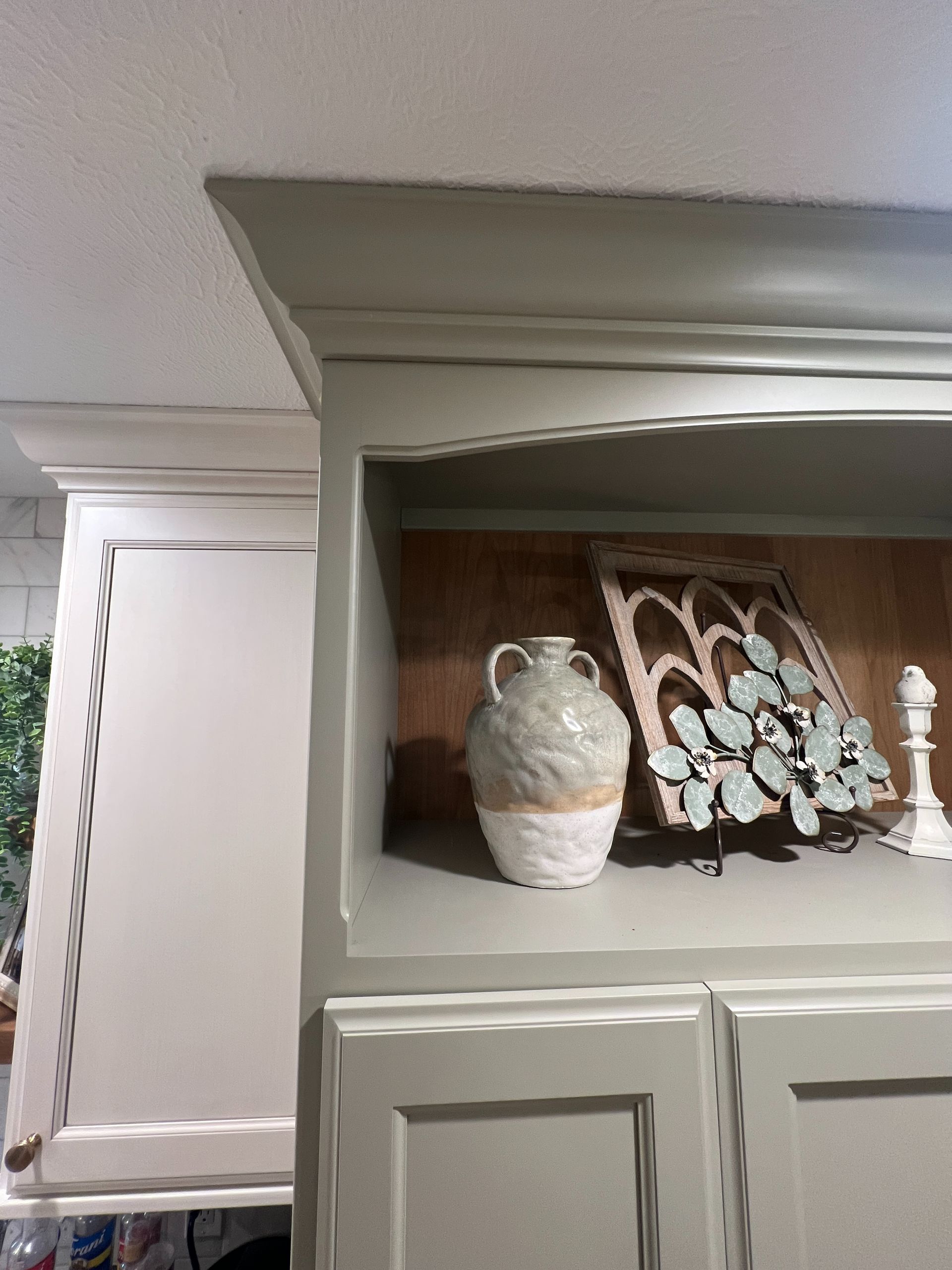 A green-grey kitchen cabinet with open shelving displays a textured vase, a decorative arched sign, and a small figurine.
