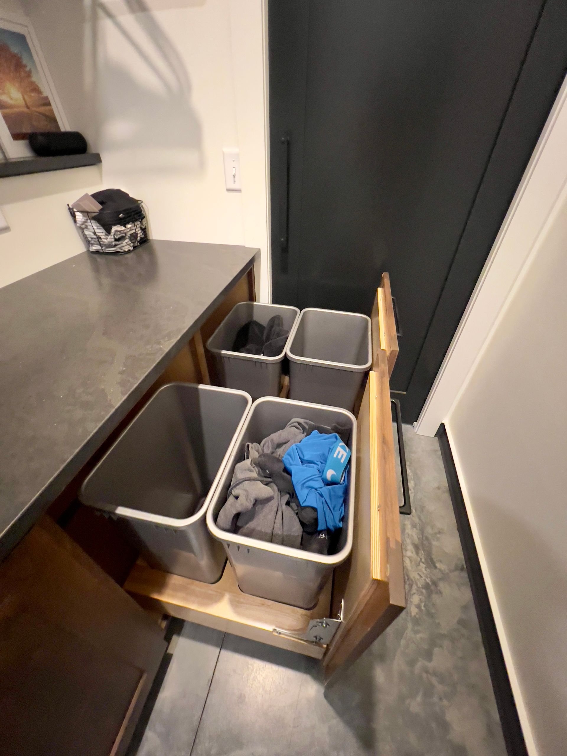A pull-out cabinet drawer in a laundry room containing four gray bins, some holding clothes, under a gray countertop.