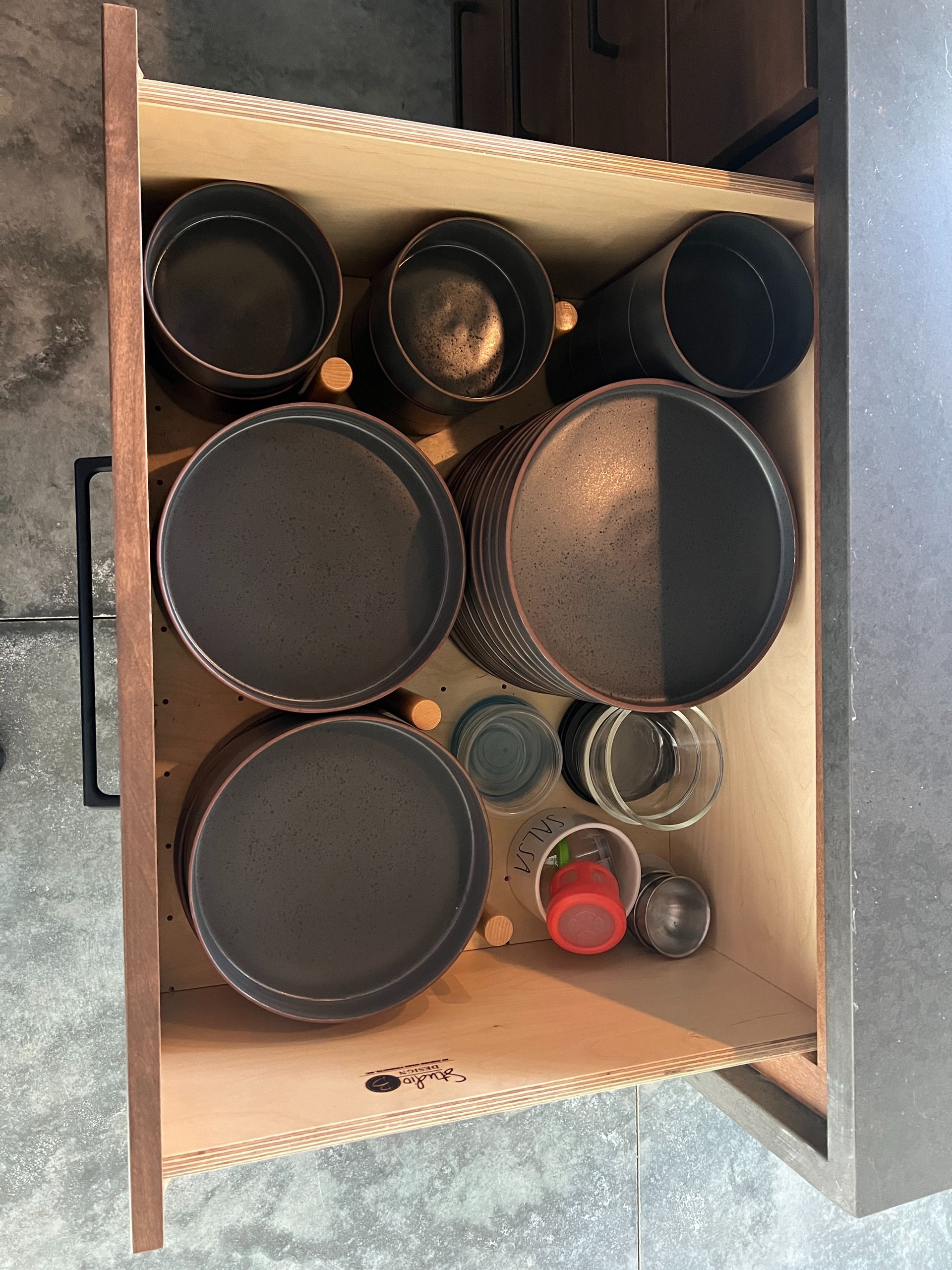 An open drawer containing several dark, circular ceramic dishes, bowls, and smaller miscellaneous items on wooden pegs.