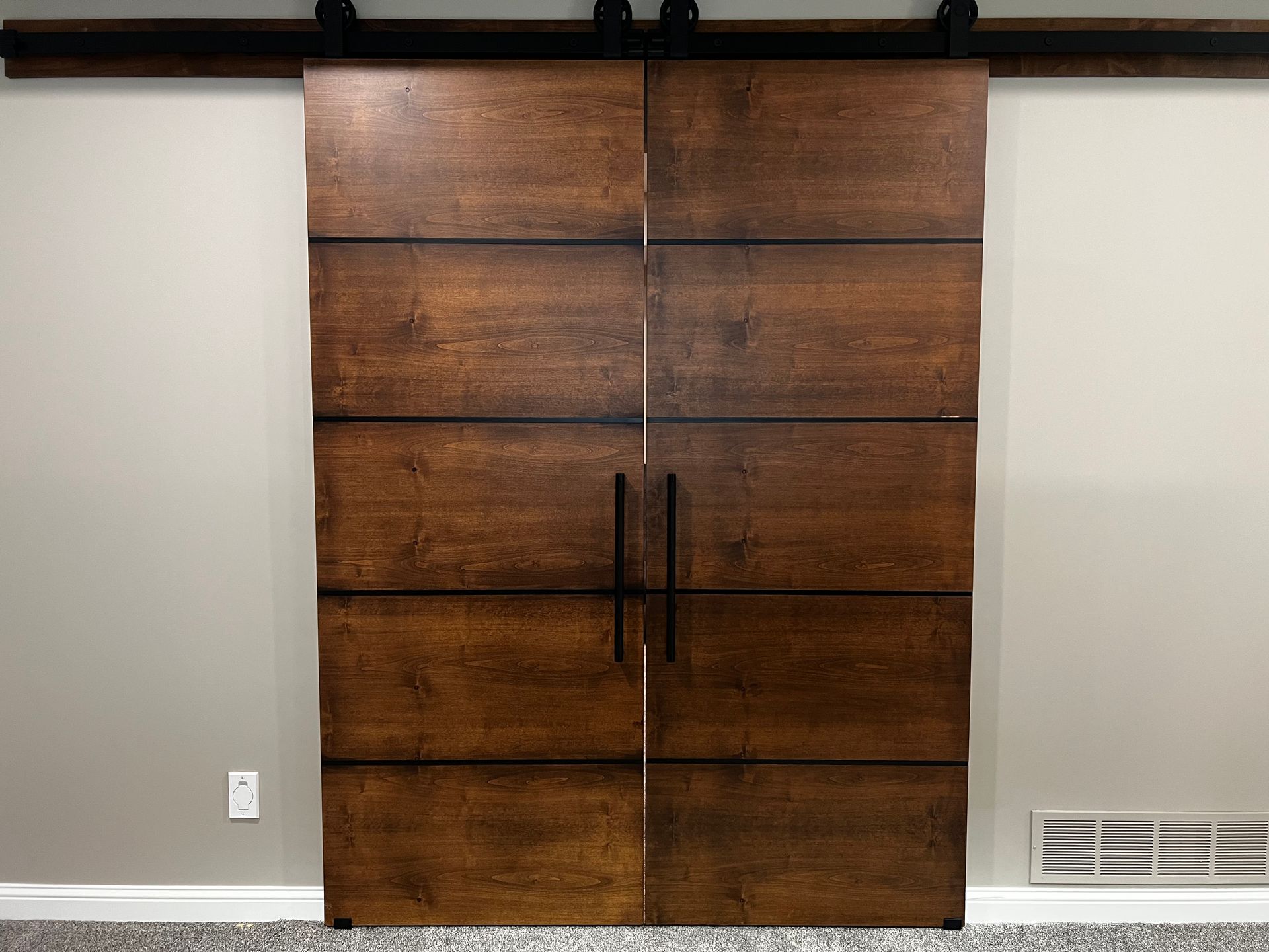 A pair of dark wood double barn doors with horizontal black metal accents, mounted on a black rail against a beige wall.