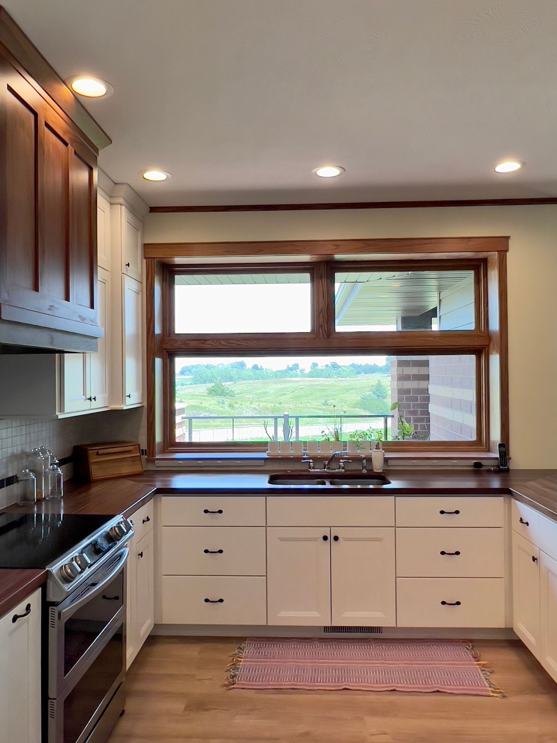 A kitchen with white cabinets, dark wood countertops, a stainless steel stove, and a large window overlooking a field.