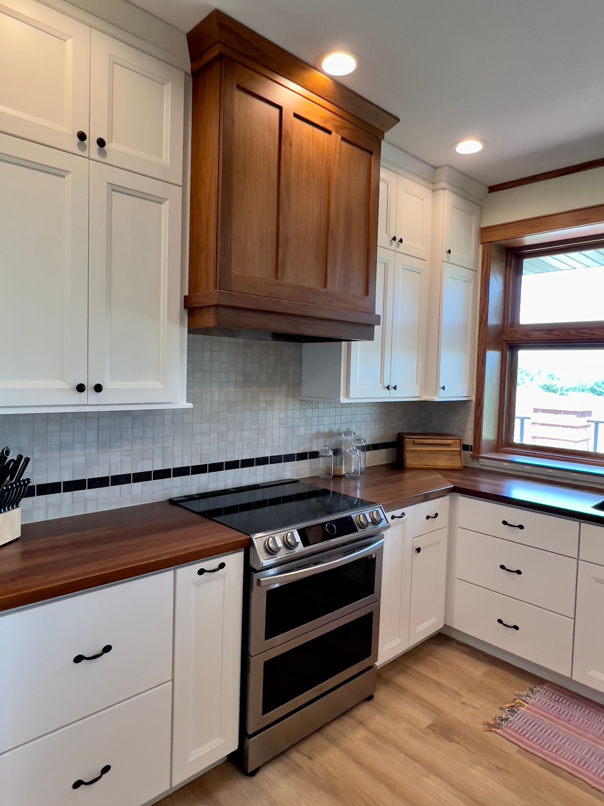 A kitchen with white cabinets, wood countertops, a custom wood vent hood over a stainless-steel range, and a window.