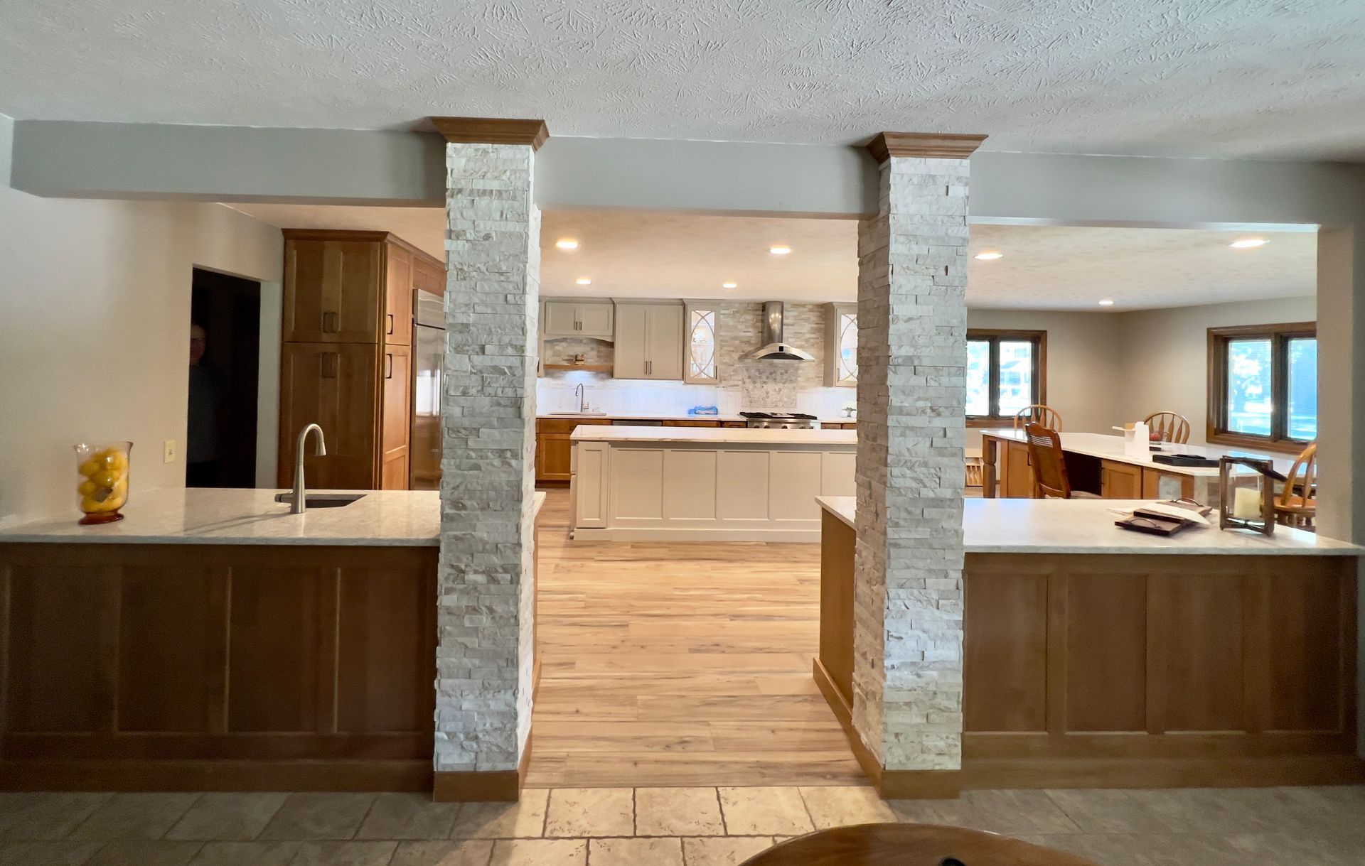 A kitchen features two wood-paneled islands separated by a central walkway flanked by two stone-faced pillars.
