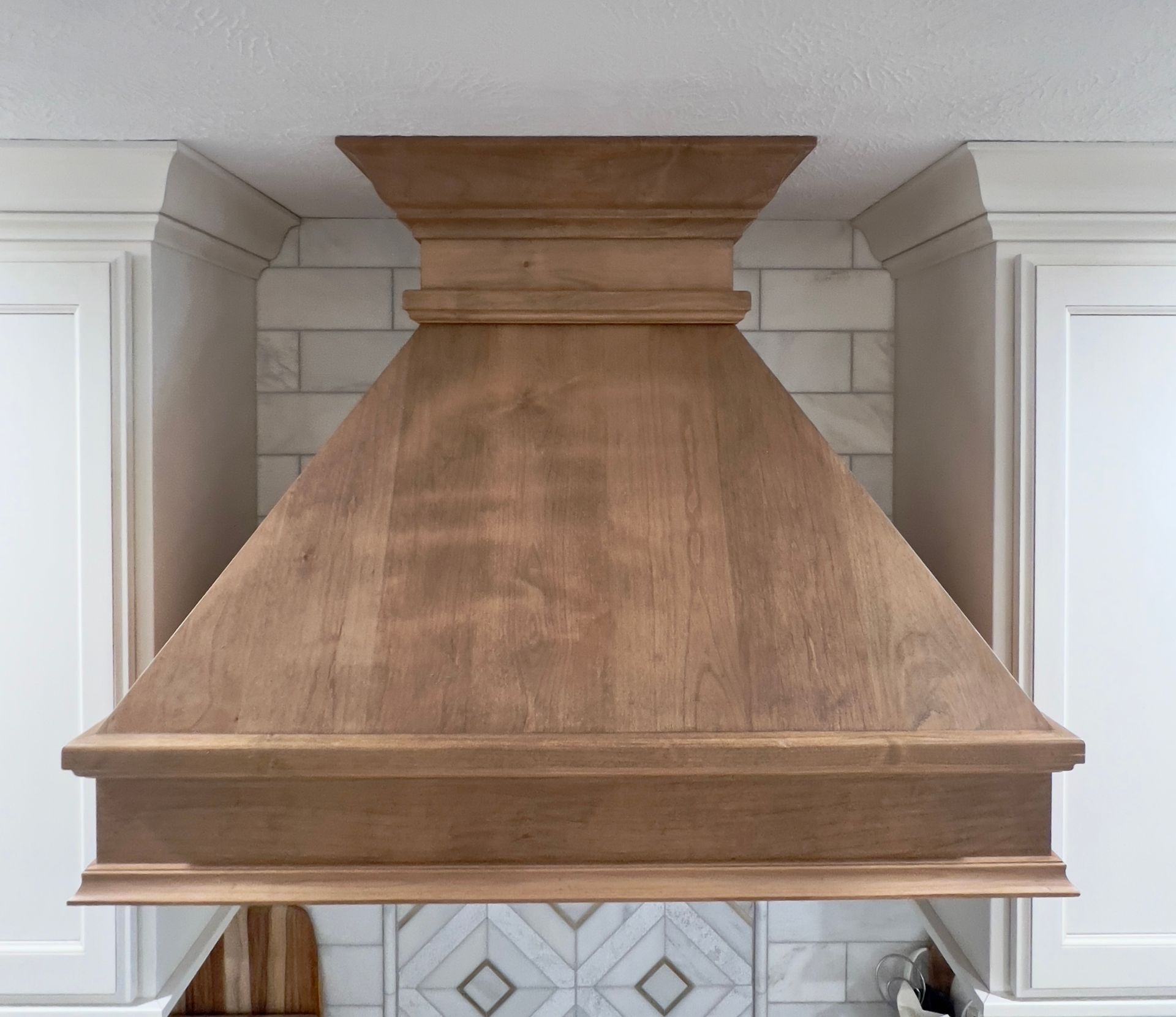 A wood range hood with crown molding centered between white kitchen cabinets against a tiled backsplash.