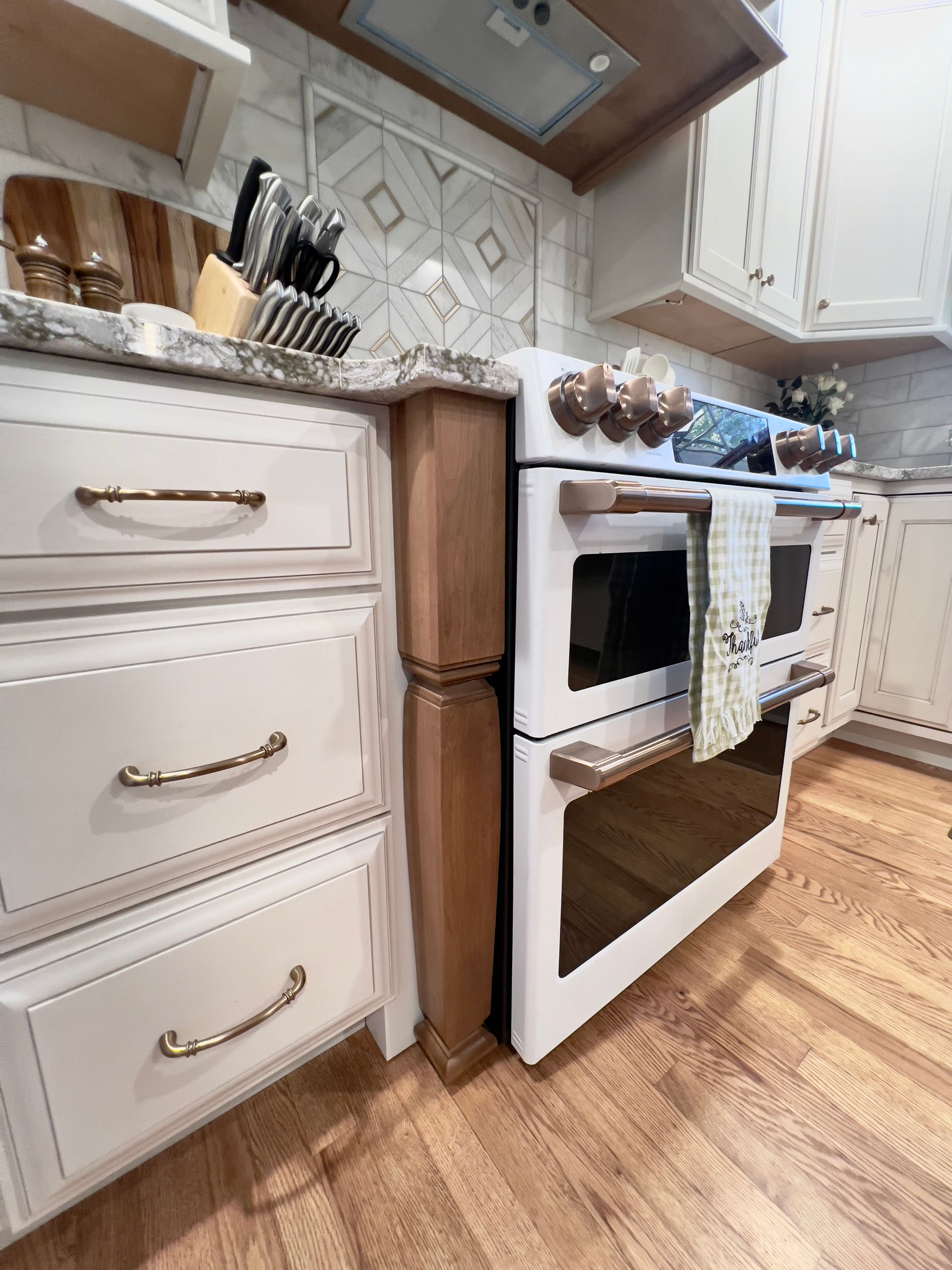 White drawers with gold hardware next to a rustic wooden column and a white double oven in a kitchen.