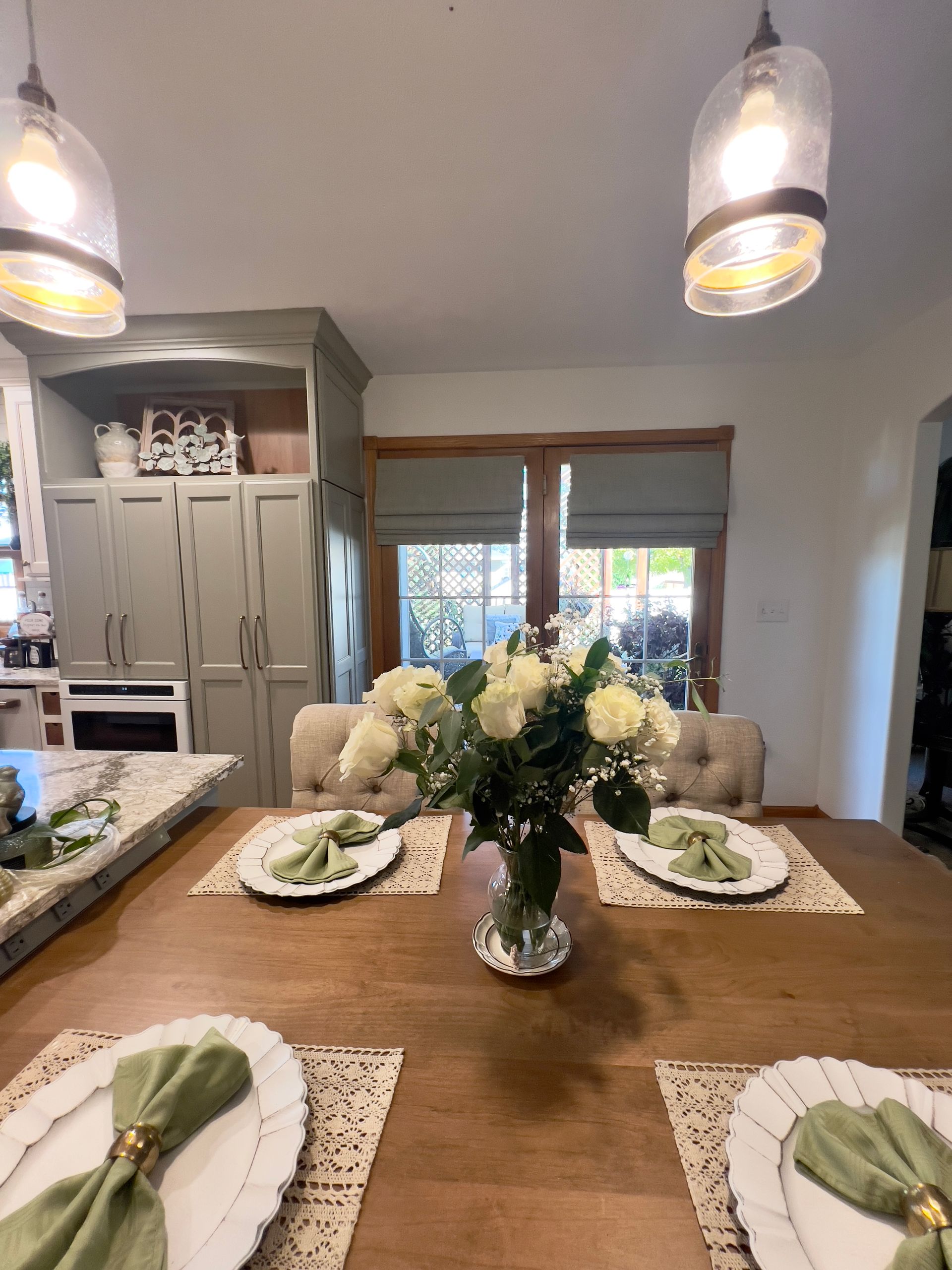 A dining room table set for four with white plates, green napkins, and a central vase of white roses under pendant lights.