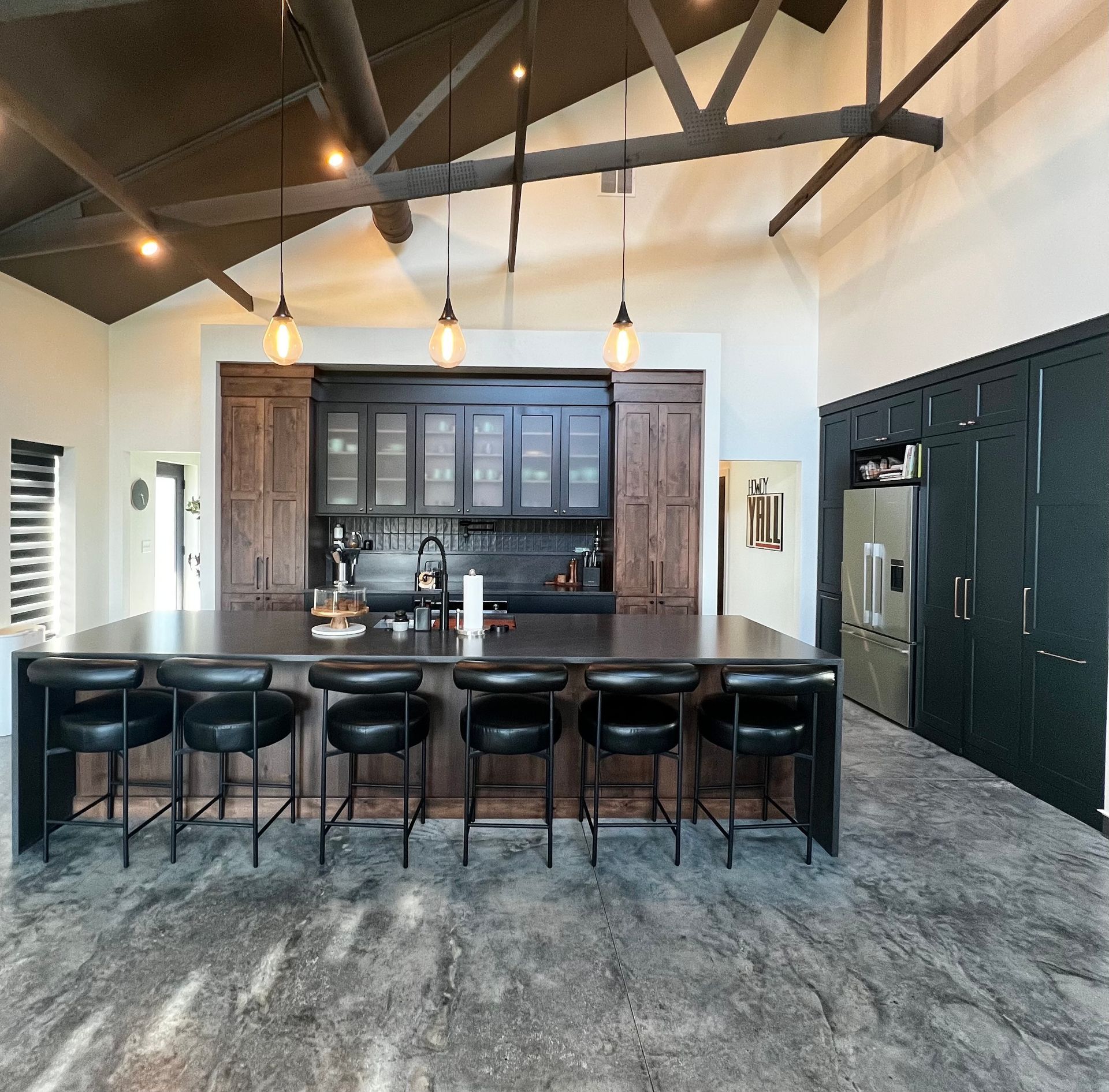 A modern kitchen features a large, dark-topped island with six black stools, dark cabinetry, and high, vaulted ceilings.