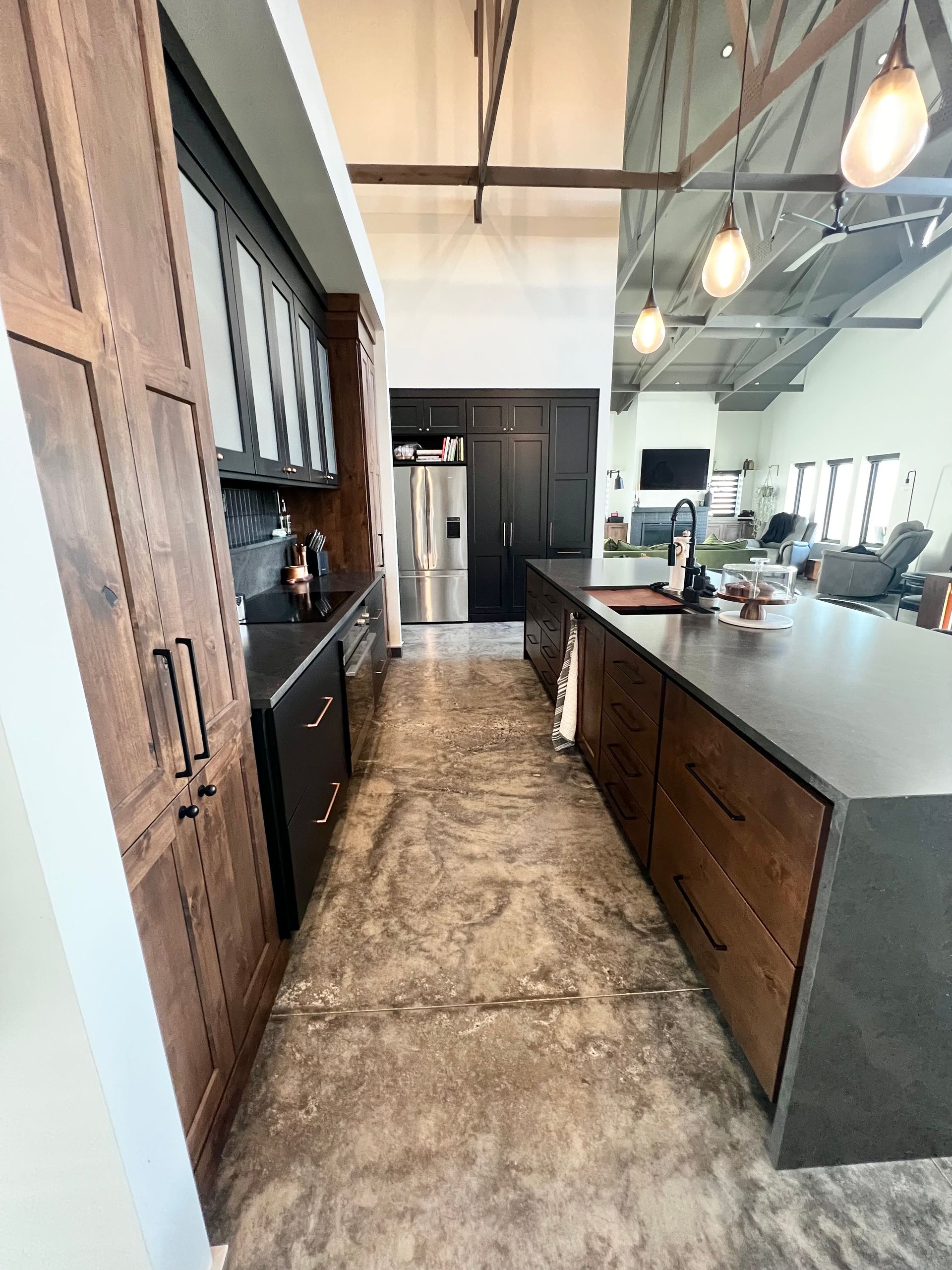 A modern kitchen with dark cabinetry, a large kitchen island, and a textured concrete floor under a high, vaulted ceiling.