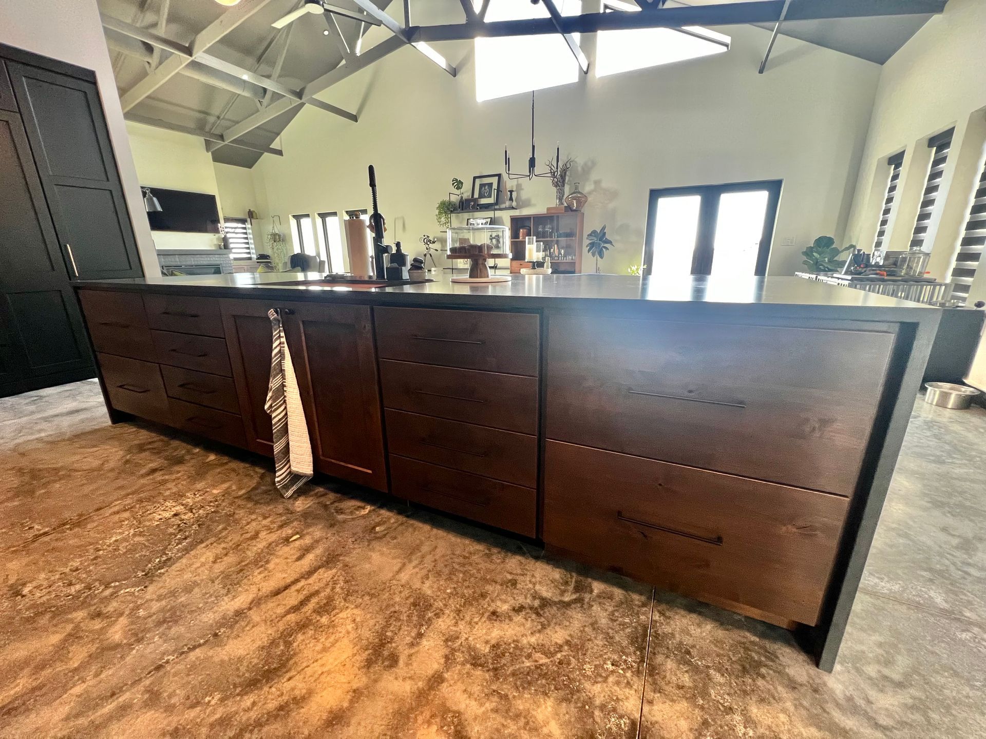 A large, dark wood kitchen island with multiple drawers sits on a concrete floor in a bright, open-plan home interior.