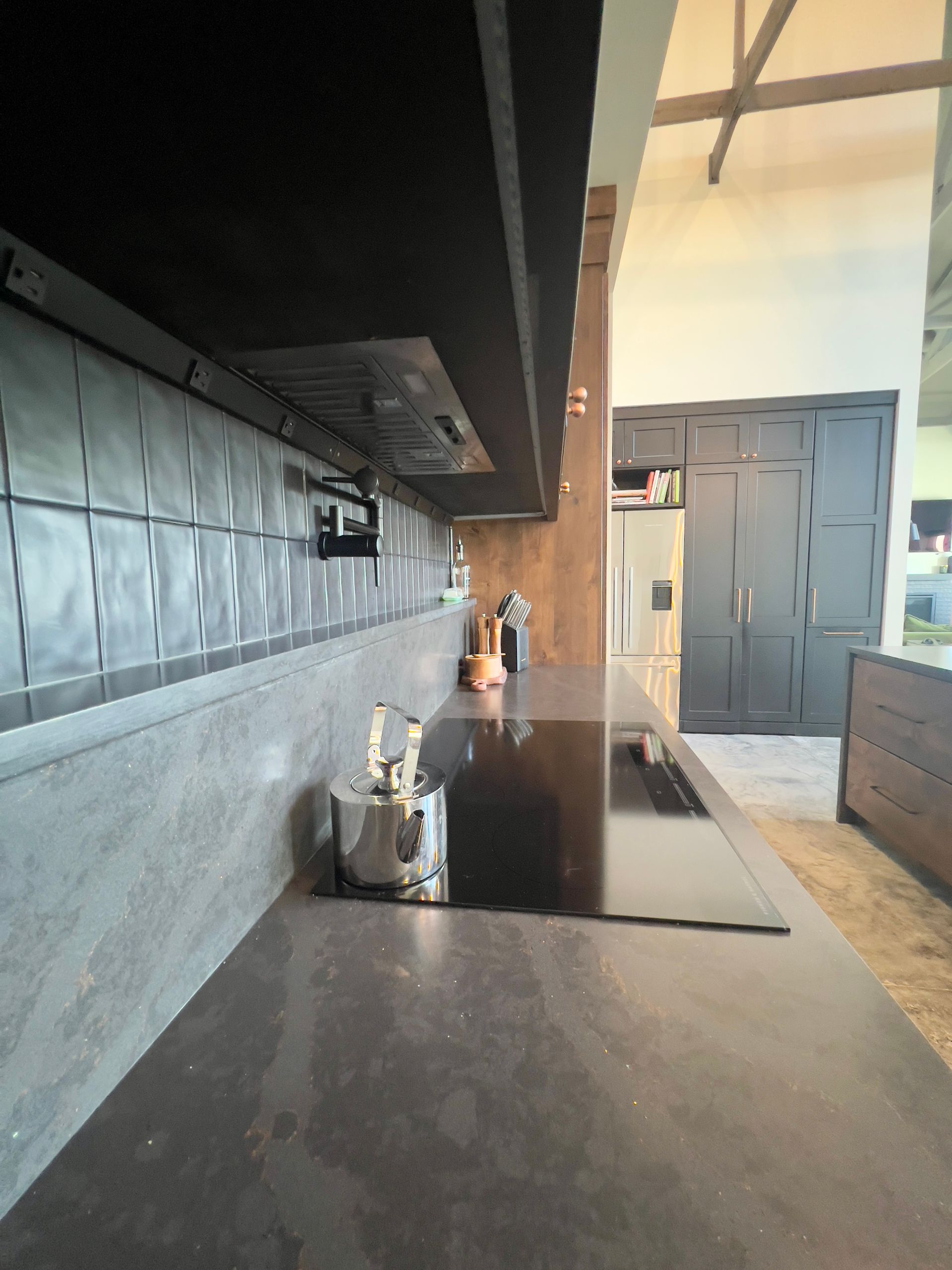 A sleek, dark-themed kitchen interior featuring a matte black countertop, induction cooktop, and gray tiled backsplash.
