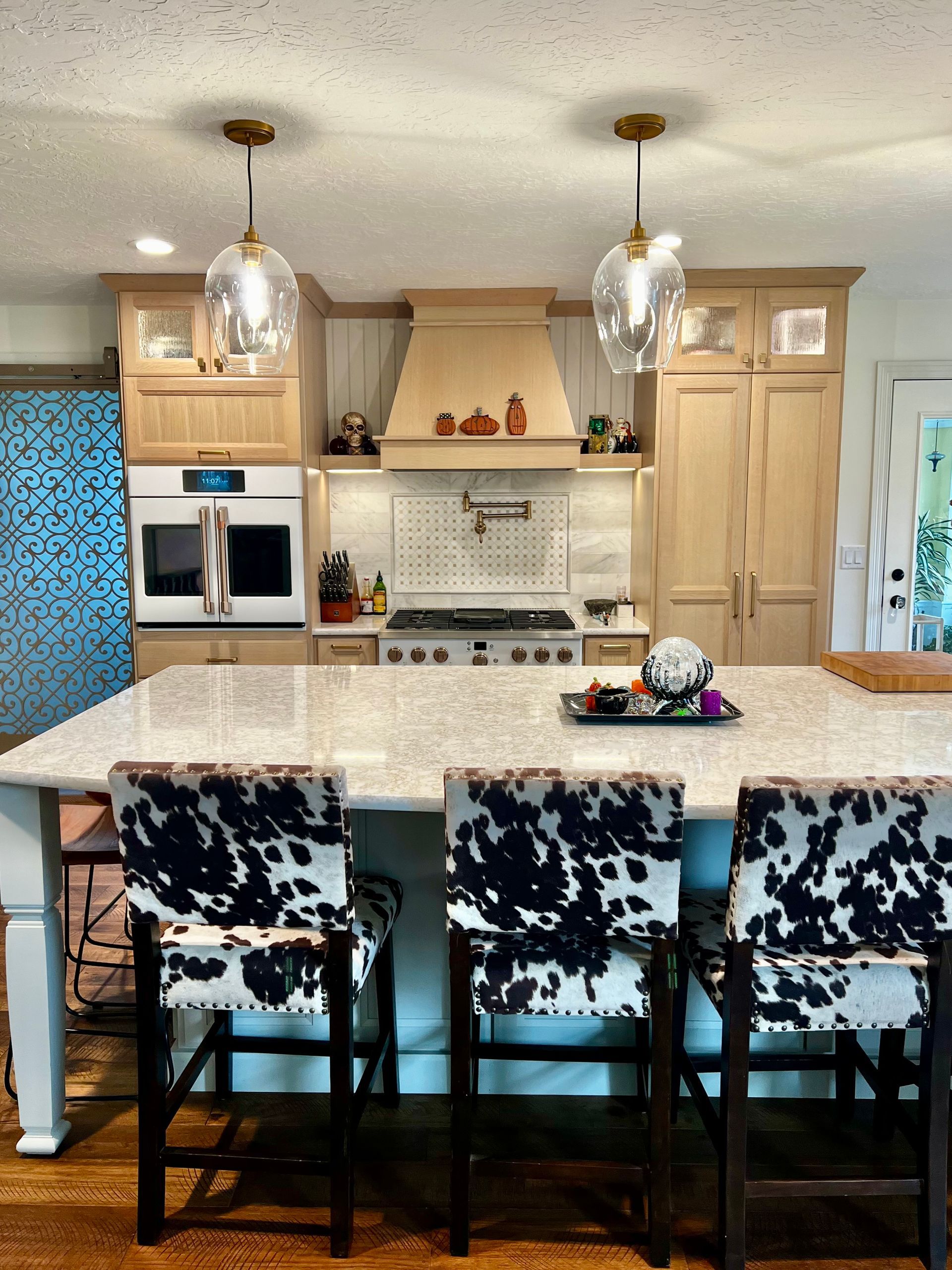 A kitchen island with three cow-print barstools sits before light wood cabinets, a range, and two hanging glass pendants.
