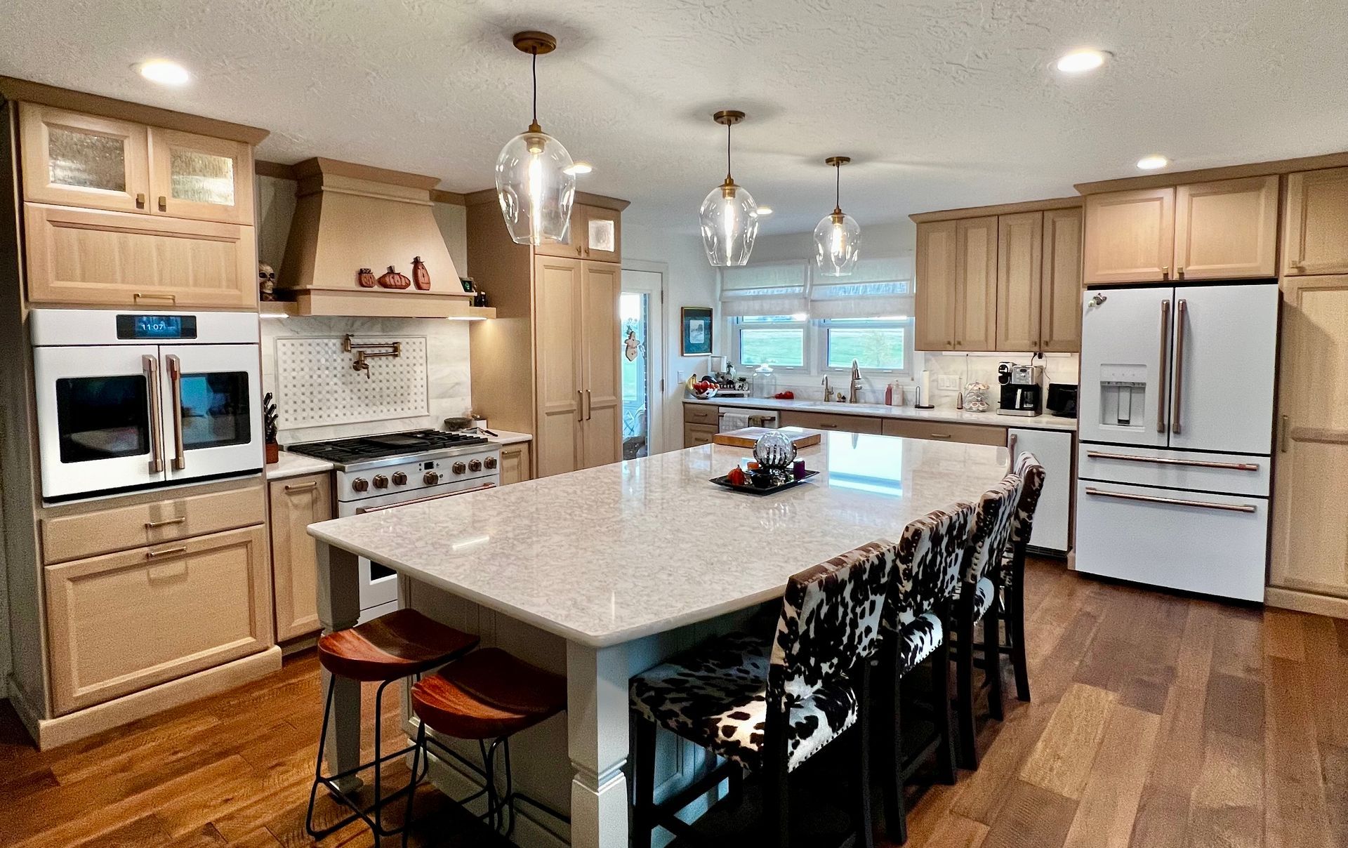 A bright kitchen with light wood cabinets, a large granite island, white appliances, and cowhide bar stools on wood floors.
