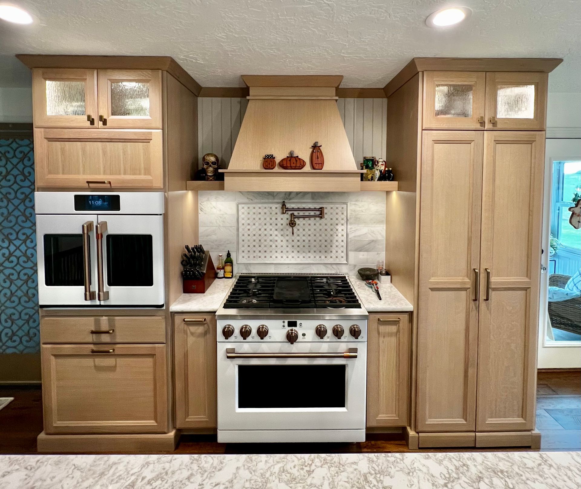 A kitchen wall with light wood cabinets, a double wall oven, a gas range with a vent hood, and a tall pantry unit.